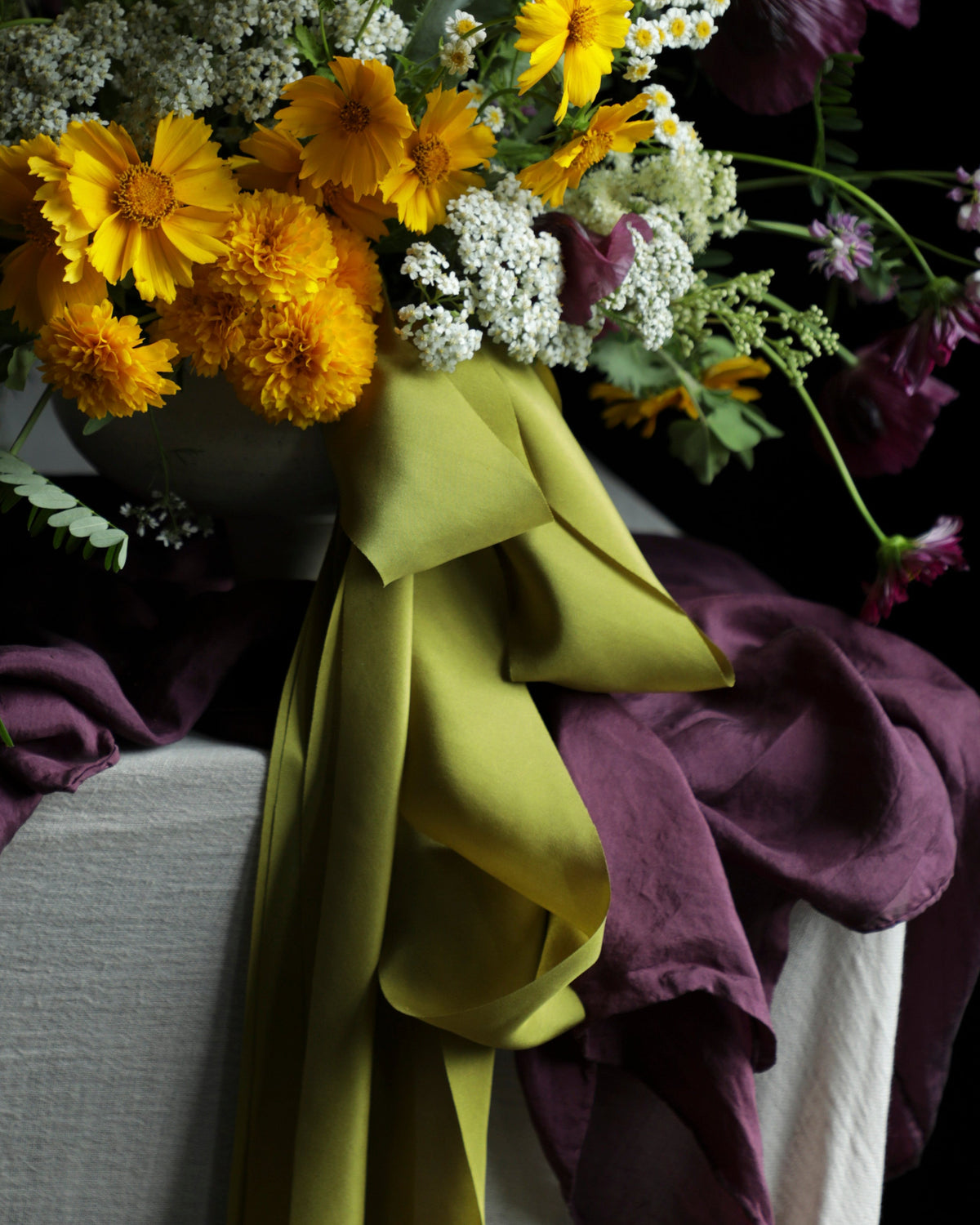 A rustic wildflower bouquet in a dark vase, featuring yellow daisies, purple poppies, white baby's breath, and a large olive green ribbon bow, set against a dark background with draped purple fabric.
