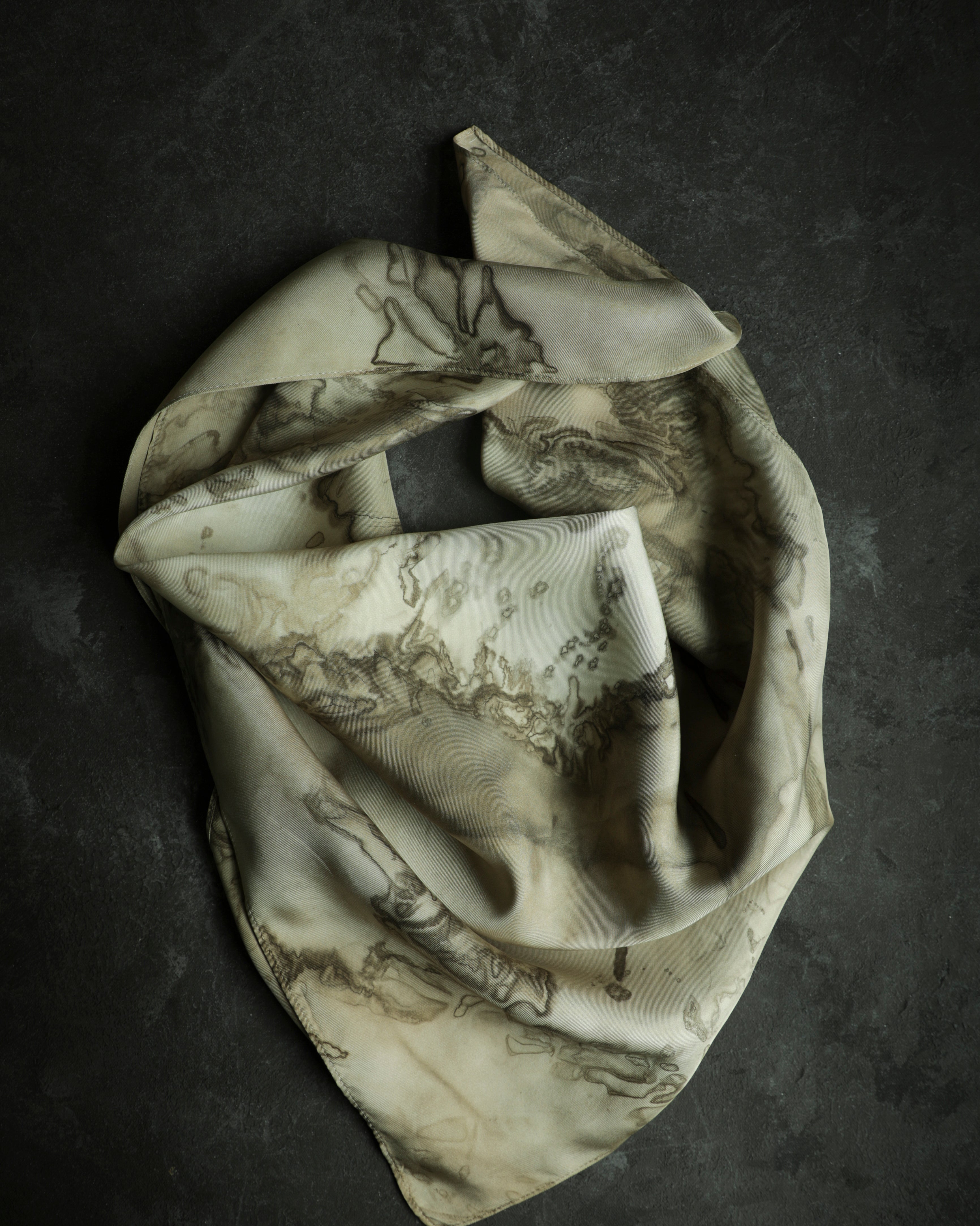 A silk twill scarf with a woodgrain-like pattern in shades of cream, champagne, and dark brown, draped and displayed on a textured background.