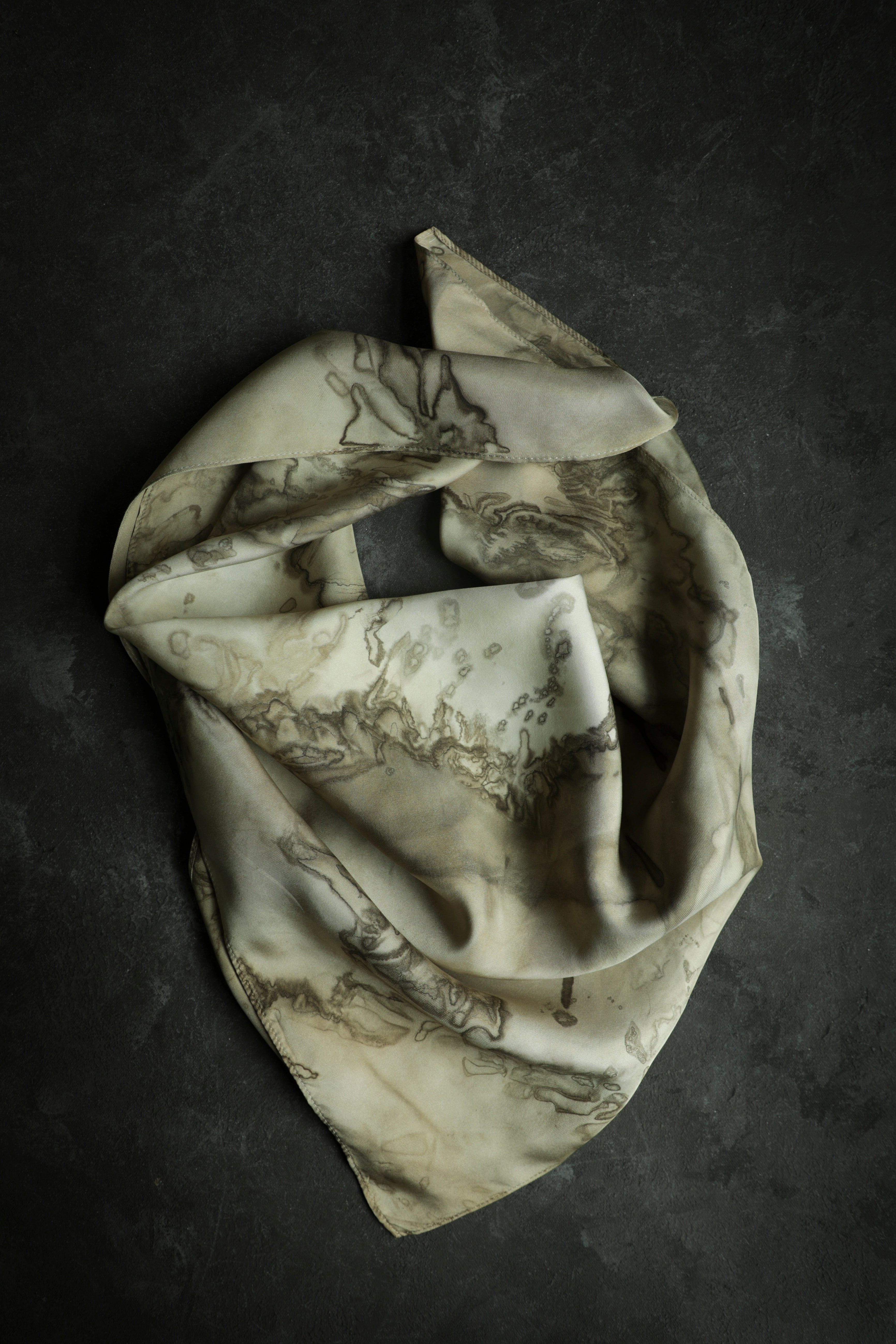 A silk twill scarf with a woodgrain-like pattern in shades of cream, champagne, and dark brown, draped and displayed on a textured background.