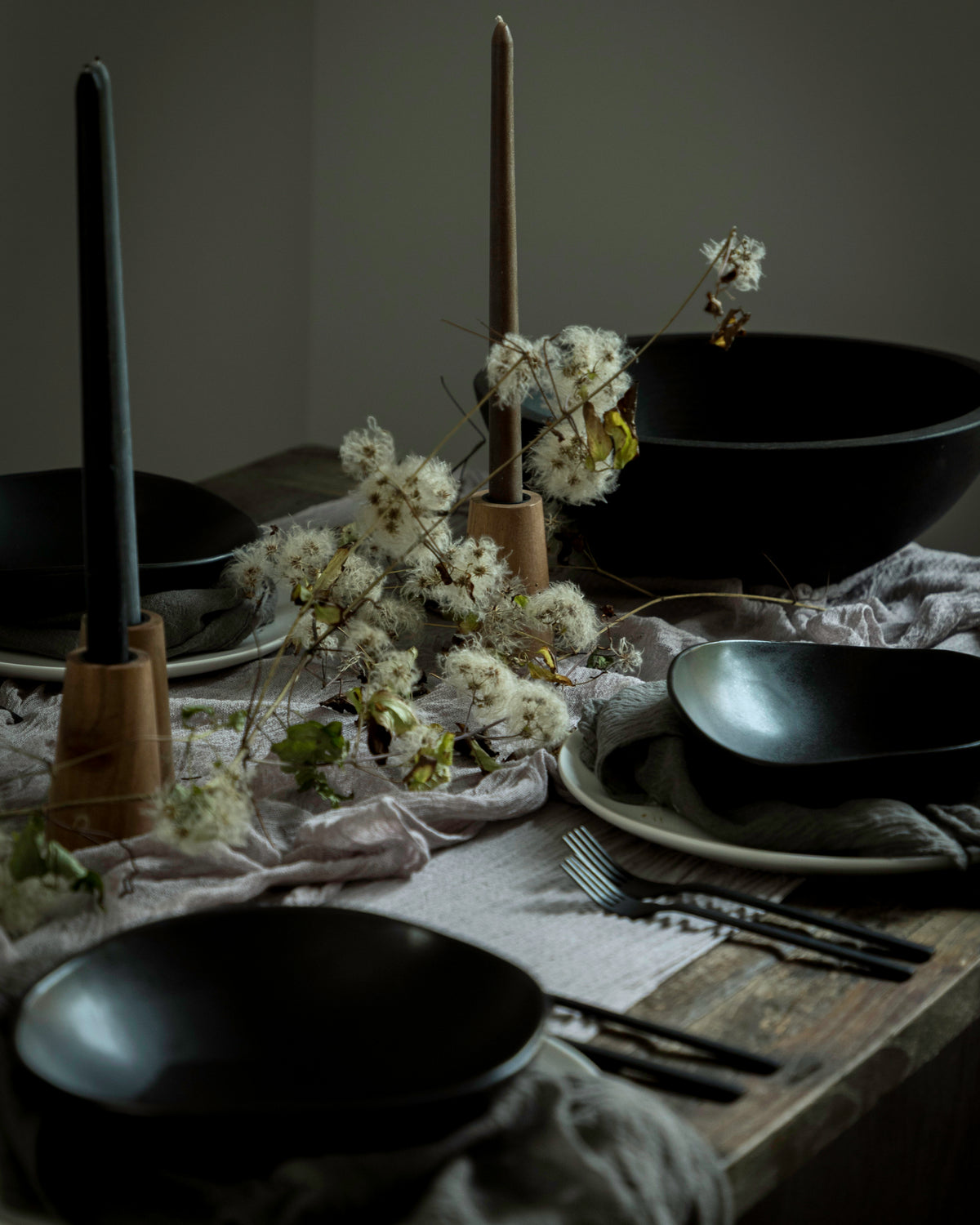 A styled dining table setting with matte black bowls, plates, black taper candles in wooden holders, and dried white flowers on a rustic wooden table with draped linen.