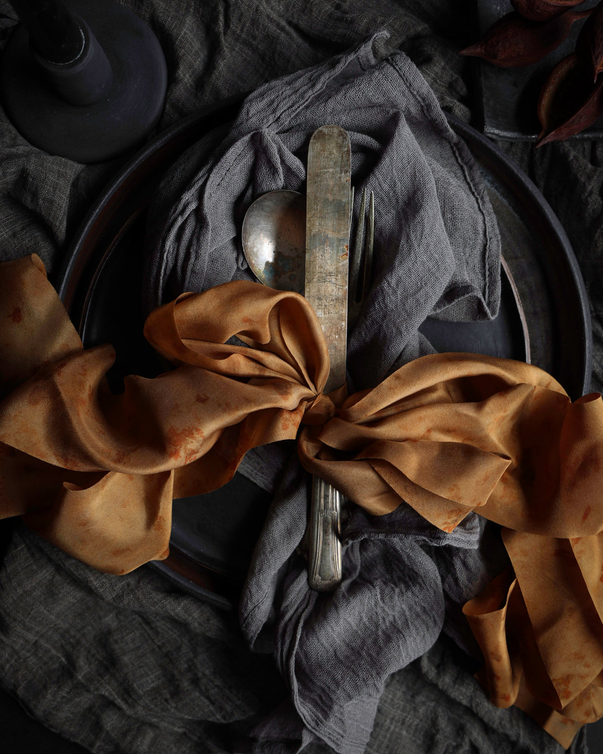 A styled tabletop setting with a dark gray linen napkin tied in a large bow with rust-brown fabric, a silver knife, and a spoon on a dark plate over a textured dark gray cloth.