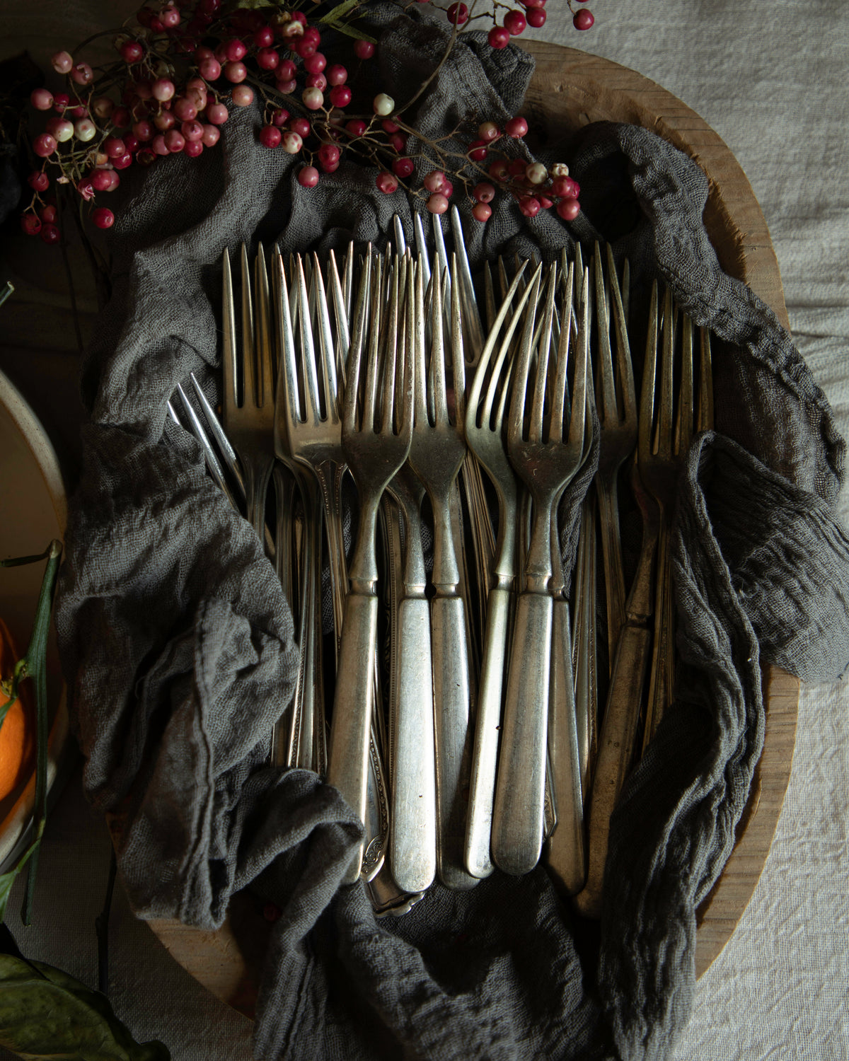 A set of vintage silver forks arranged in a wooden bowl draped with a dark gray cloth, decorated with red berries on a neutral fabric background.