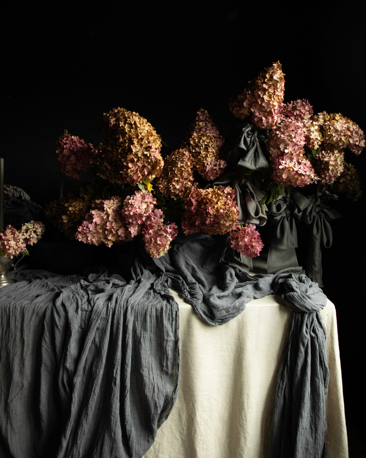 A still life arrangement of dried pink and brown flowers draped over a table covered with dark gray and off-white fabric, set against a solid black background.