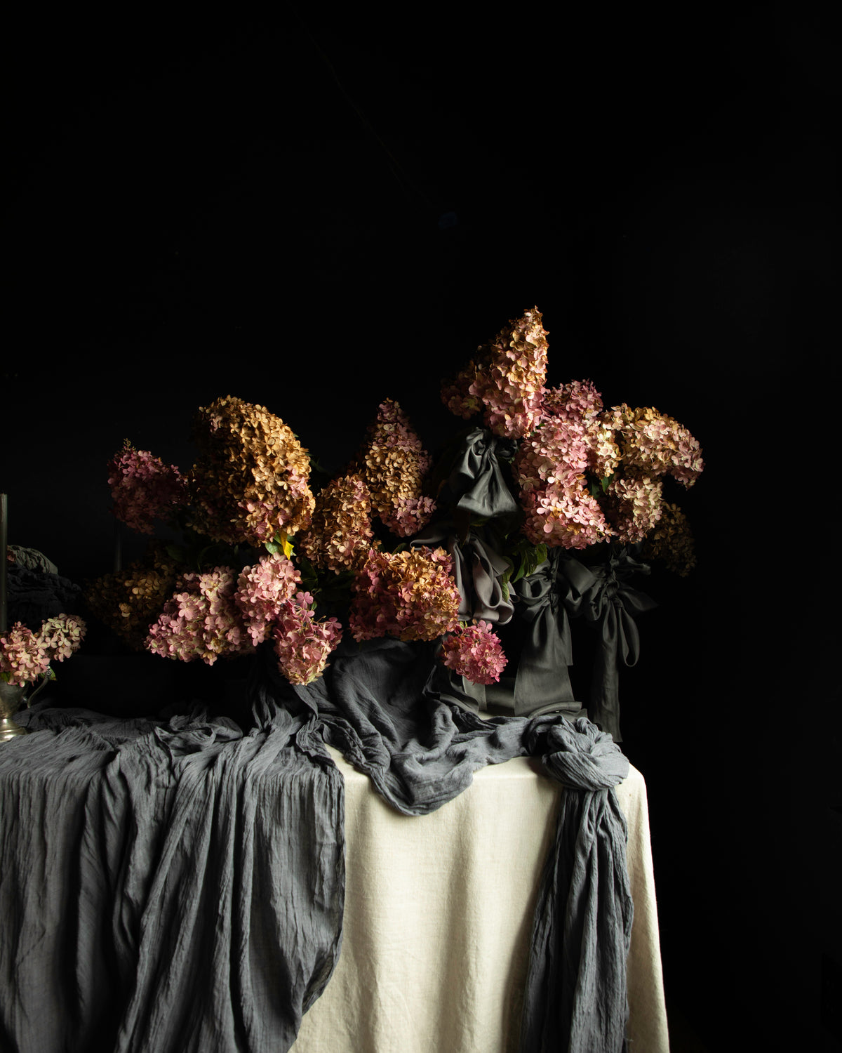A still life arrangement of dried pink and brown flowers draped over a table covered with dark gray and off-white fabric, set against a solid black background.