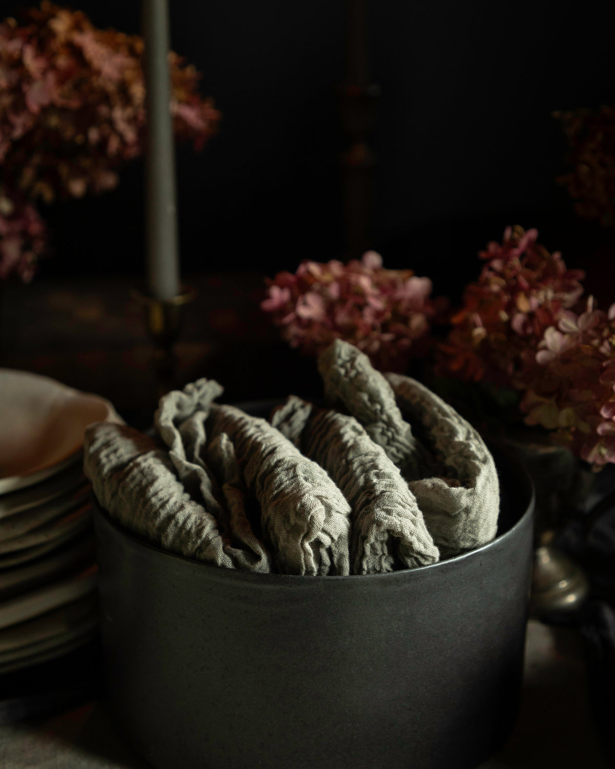 A dark round container filled with dried, wrinkled sage leaves, styled on a table with plates and pink hydrangeas in a dim, moody setting.
