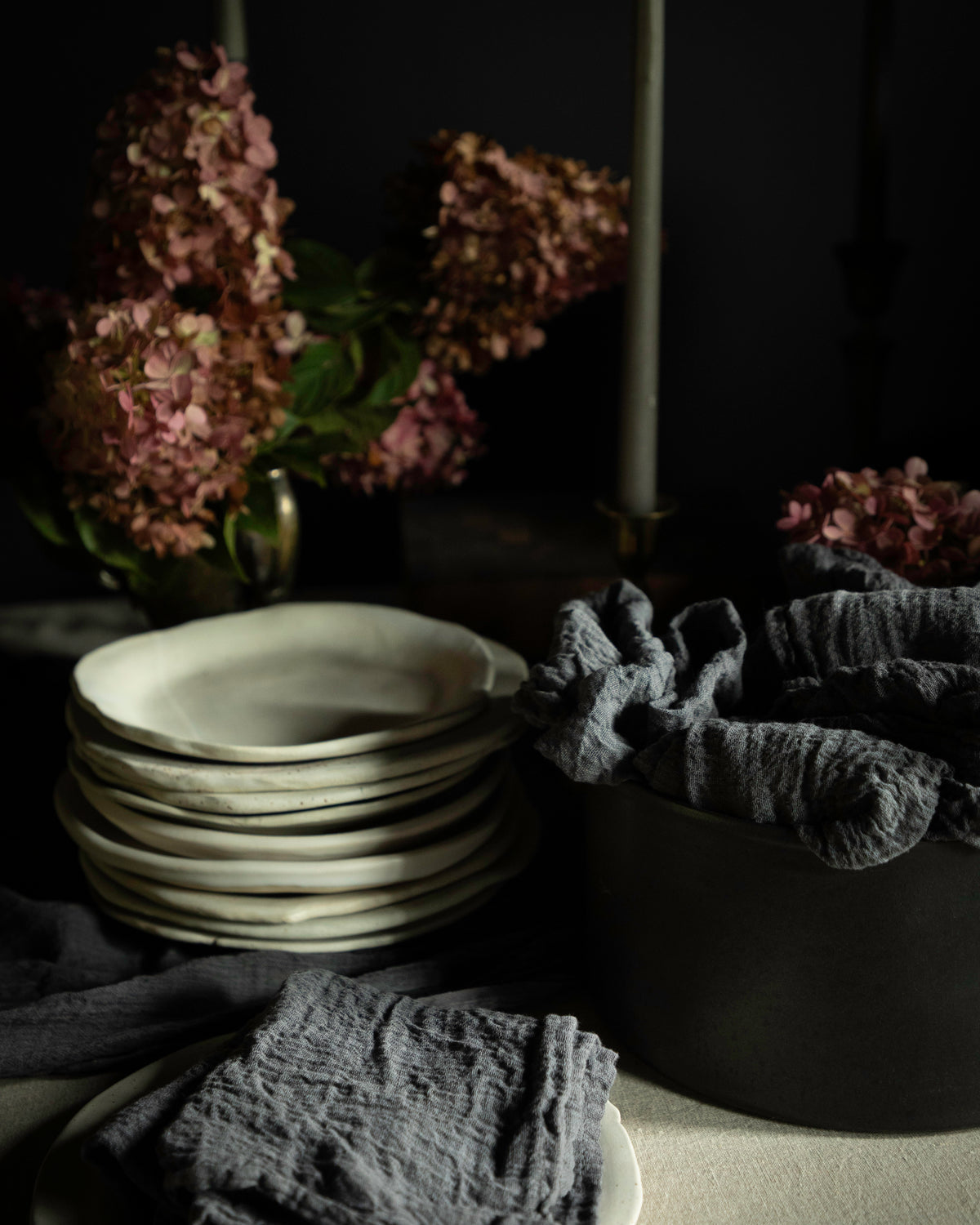 A styled tabletop scene with a stack of matte off-white ceramic plates, dark gray textured cloth napkins draped over a black bowl, and dried pink hydrangea flowers in the background against a dark backdrop.