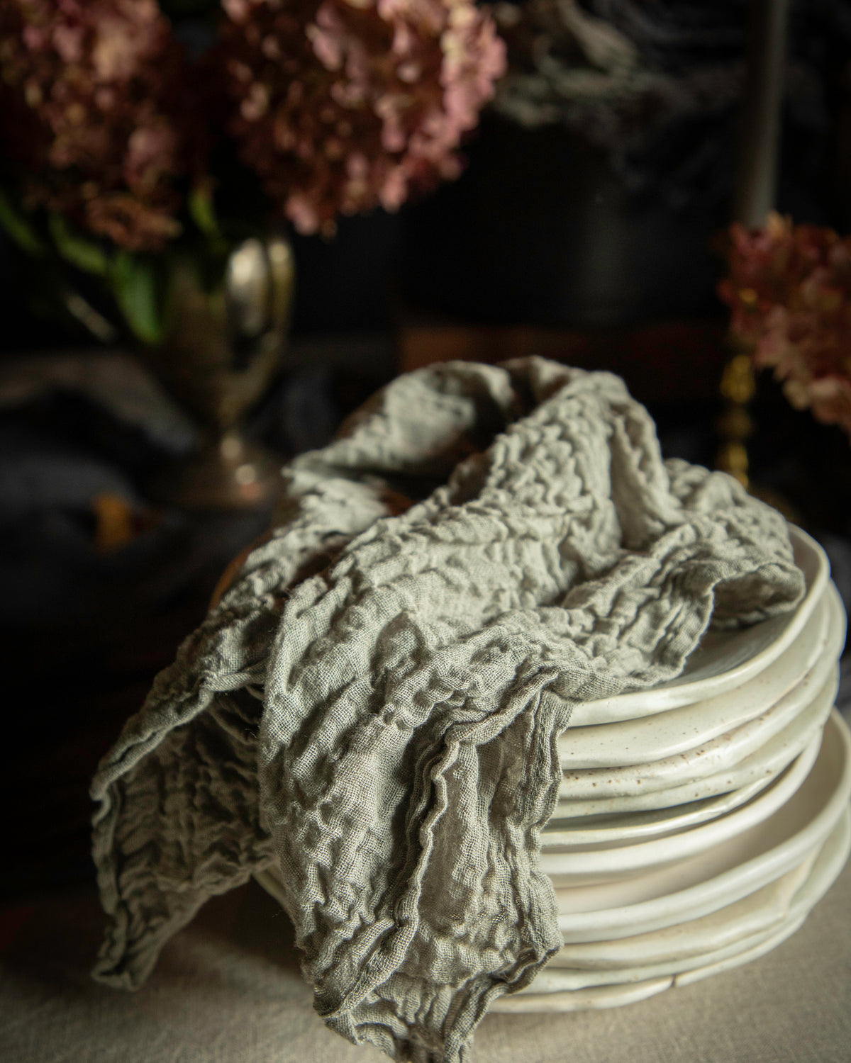 A textured, crinkled light gray linen cloth draped over a stack of white ceramic plates, set against a dark, moody background with blurred pink flowers.