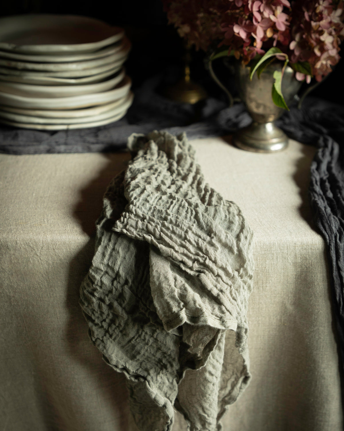 A crinkled, textured gray linen napkin draped over a beige tablecloth in a moody, rustic setting with plates and flowers in the background.