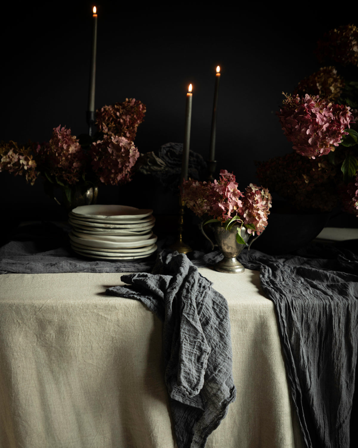 A moody tabletop still life with a cream tablecloth, dark gray crinkled napkins, stacked white plates, tall black candlesticks with lit candles, and dried pinkish-brown flowers against a black background.