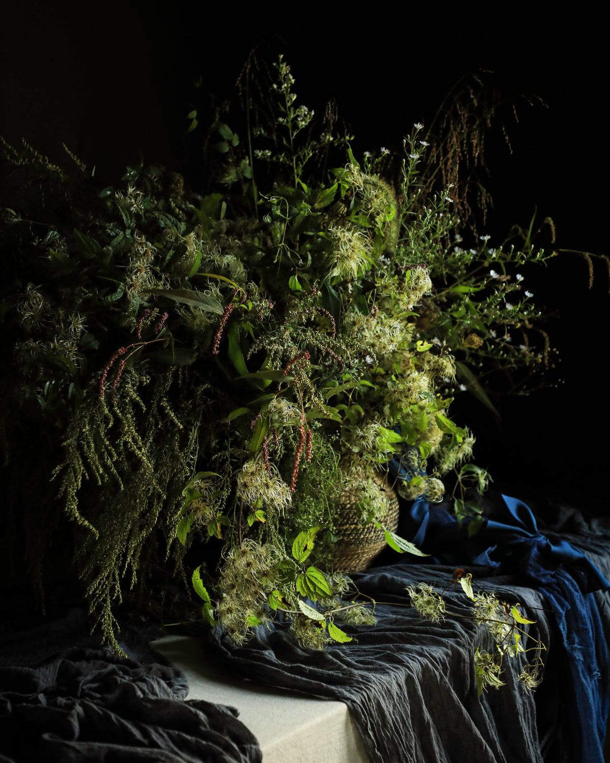 A large, rustic dried flower and botanical arrangement in earthy green and brown tones, styled on a draped dark blue and black fabric surface against a solid black background.