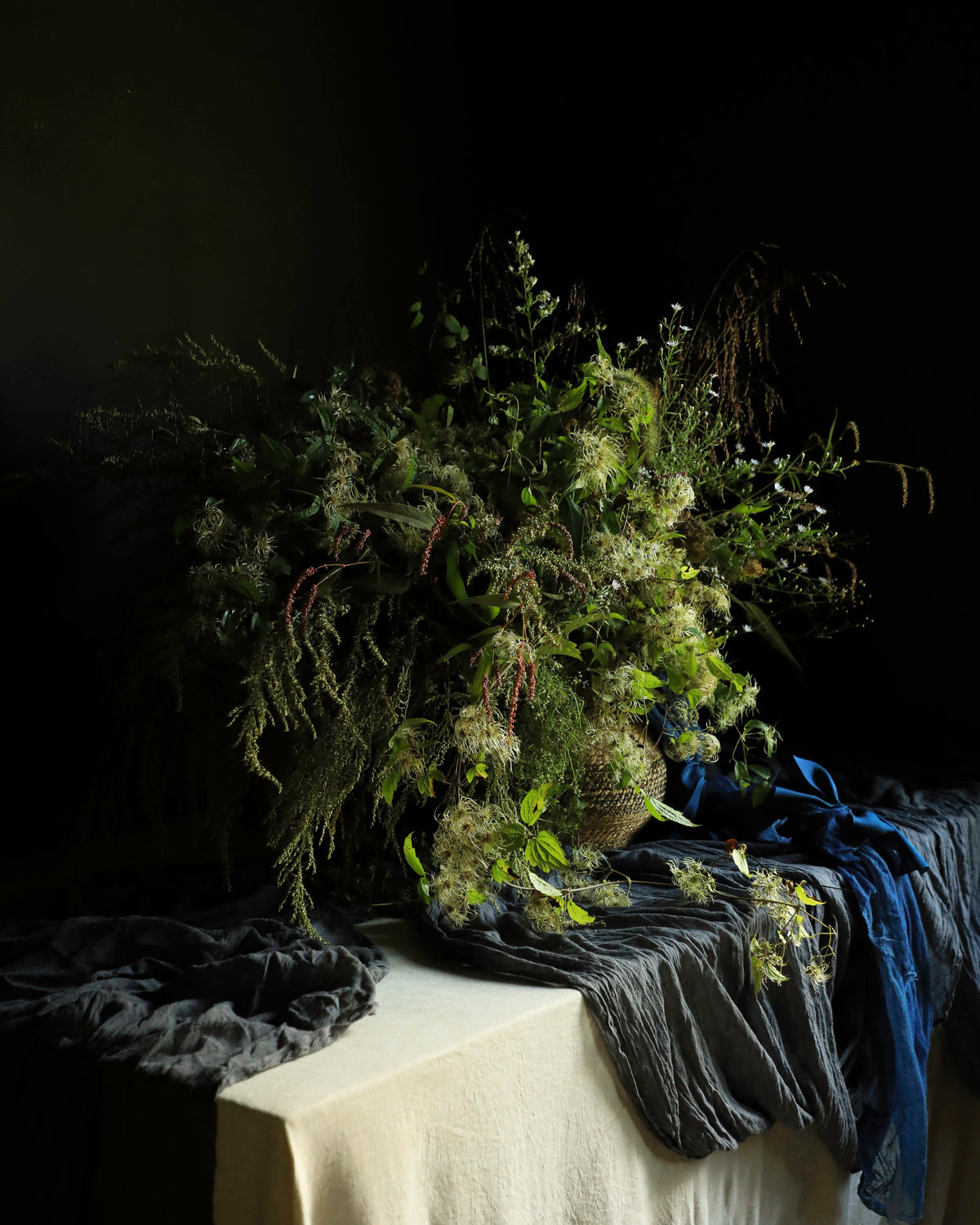 A large, rustic wildflower and foliage arrangement in a woven basket, styled on a draped table with dark gray fabric and a blue ribbon against a dark background.