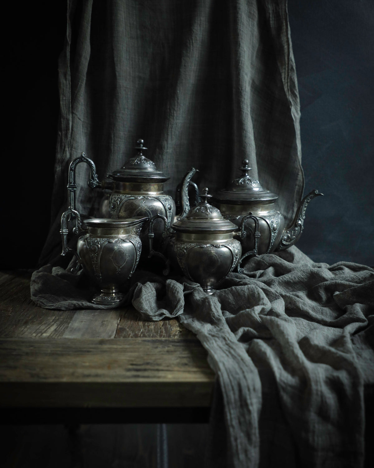 A set of ornate silver metal teapots and cups arranged on a rustic wooden table, draped with dark gray linen fabric against a dark studio background.