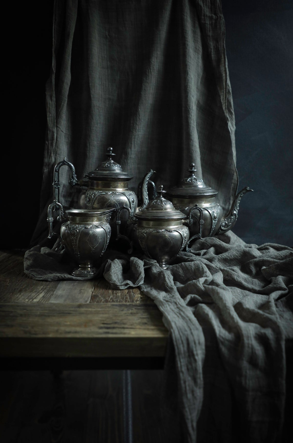 A set of ornate silver metal teapots and cups arranged on a rustic wooden table, draped with dark gray linen fabric against a dark studio background.
