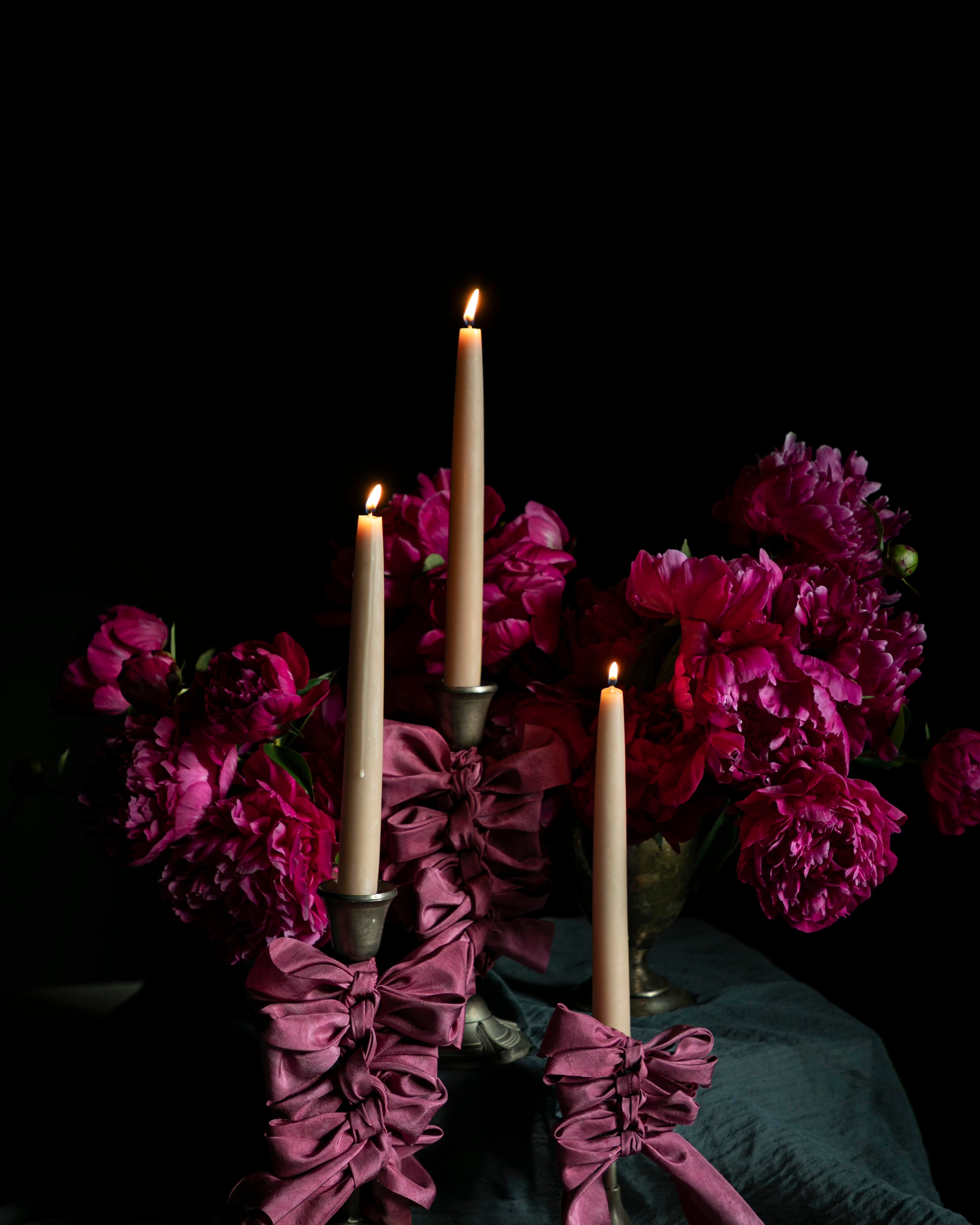 opalite aesthetic. Candle holder with purple ribbons and candles surrounded by pink peony flowers on a dark background. wedding table inspiration