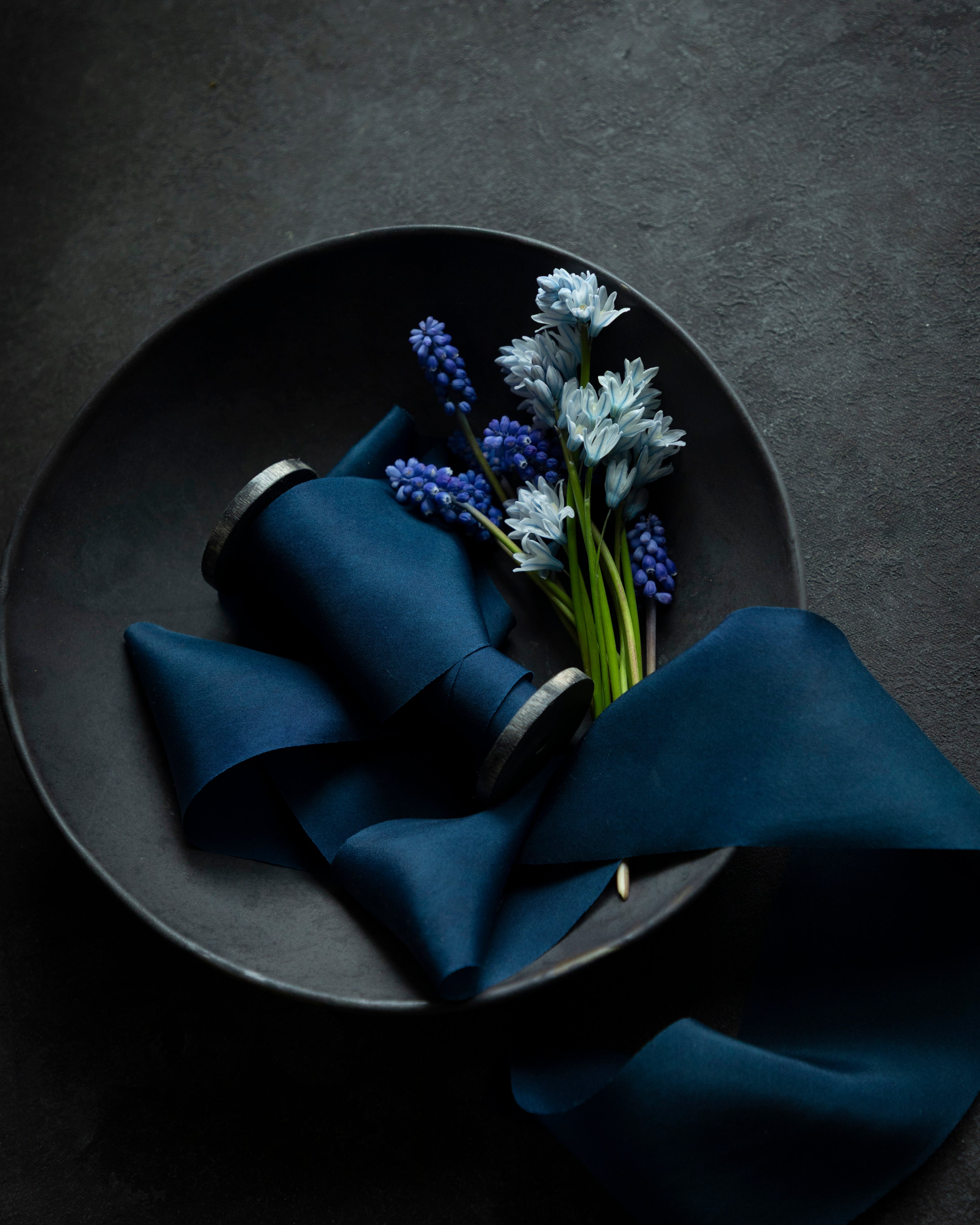 A dark blue silk ribbon displayed on a dark ceramic bowl with a small arrangement of blue and white flowers, and a sample swatch of the ribbon placed beside it.