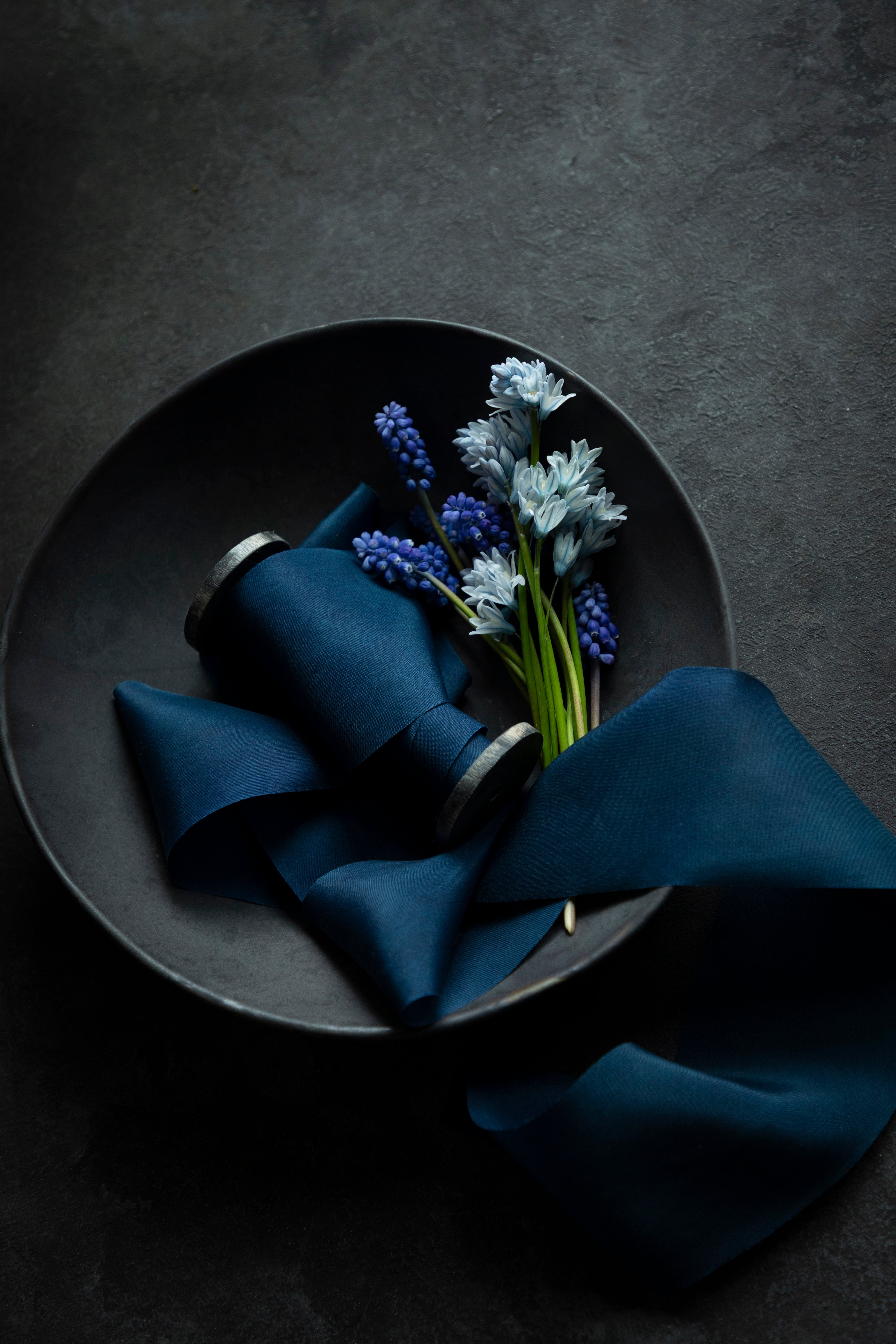 A dark blue silk ribbon displayed on a dark ceramic bowl with a small arrangement of blue and white flowers, and a sample swatch of the ribbon placed beside it.