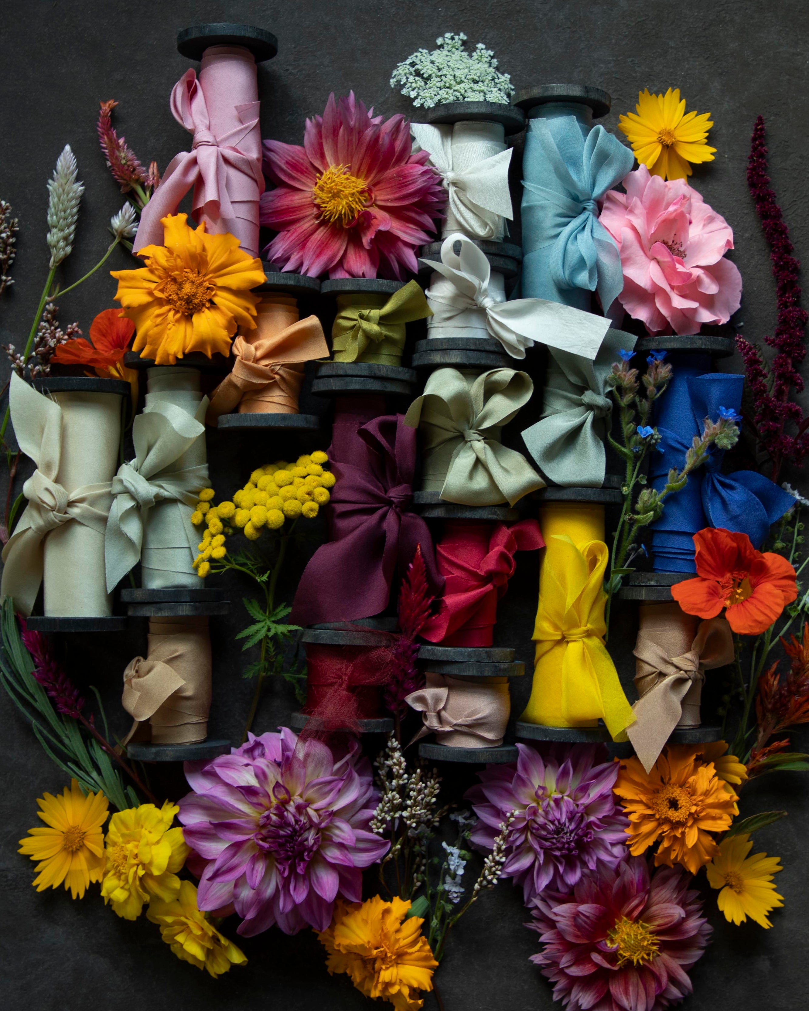 A styled flat-lay of multiple colorful silk ribbon spools wrapped with ribbons and surrounded by vibrant flowers, arranged on a dark textured background.