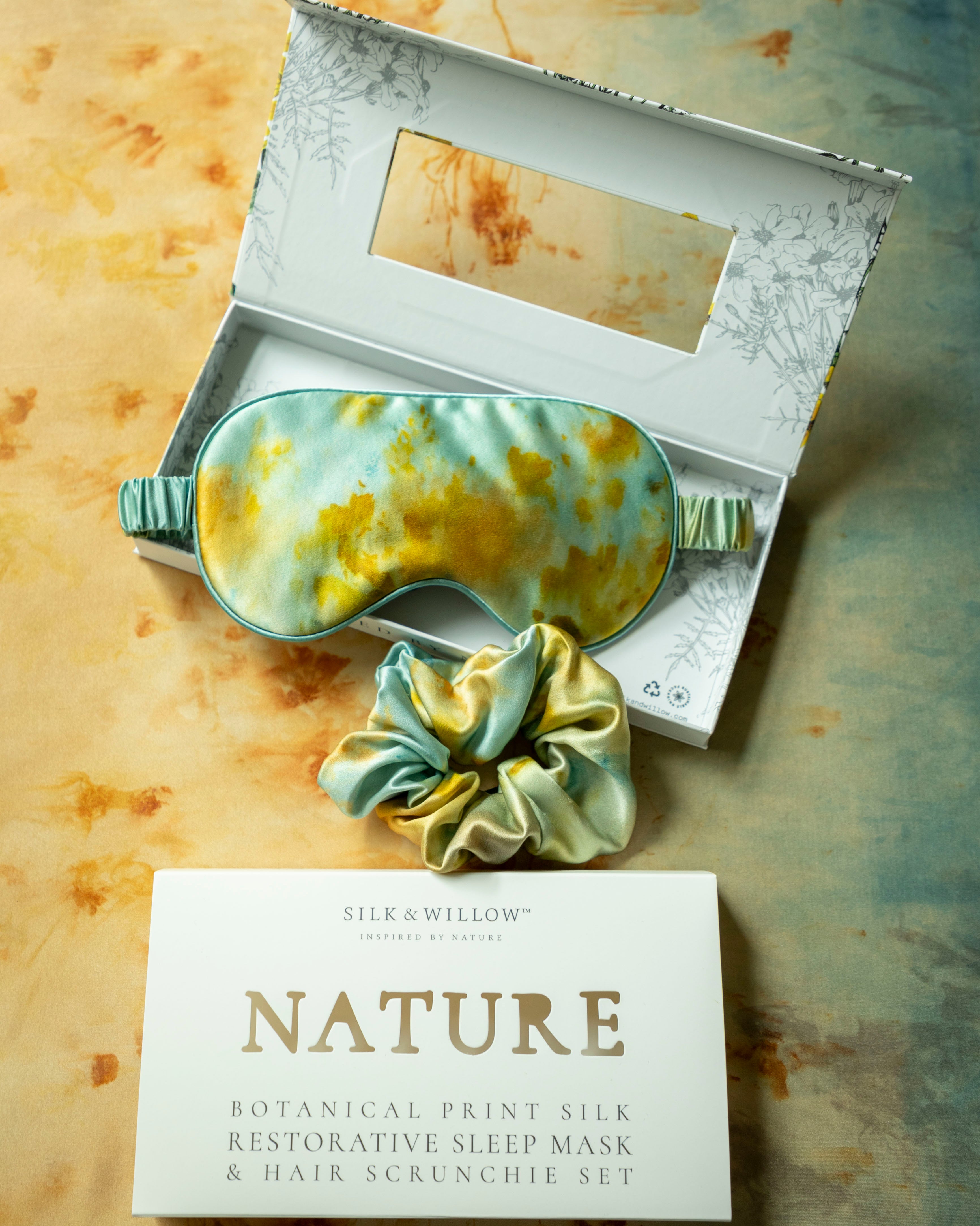 Silk & Willow Nature botanical print silk restorative sleep mask and hair scrunchie set on a textured surface.