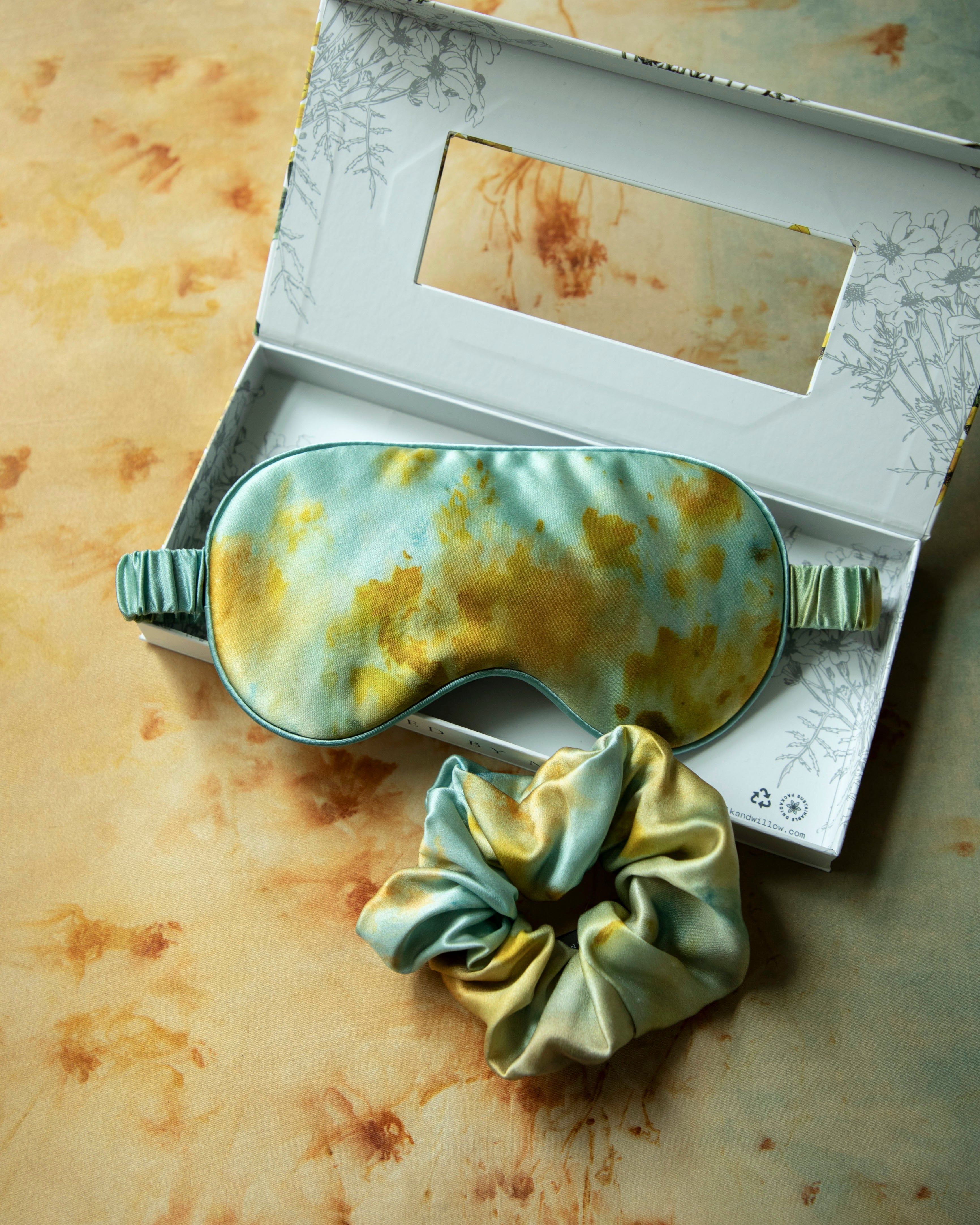 Sleep mask and scrunchie set with vintage-style packaging on a textured surface