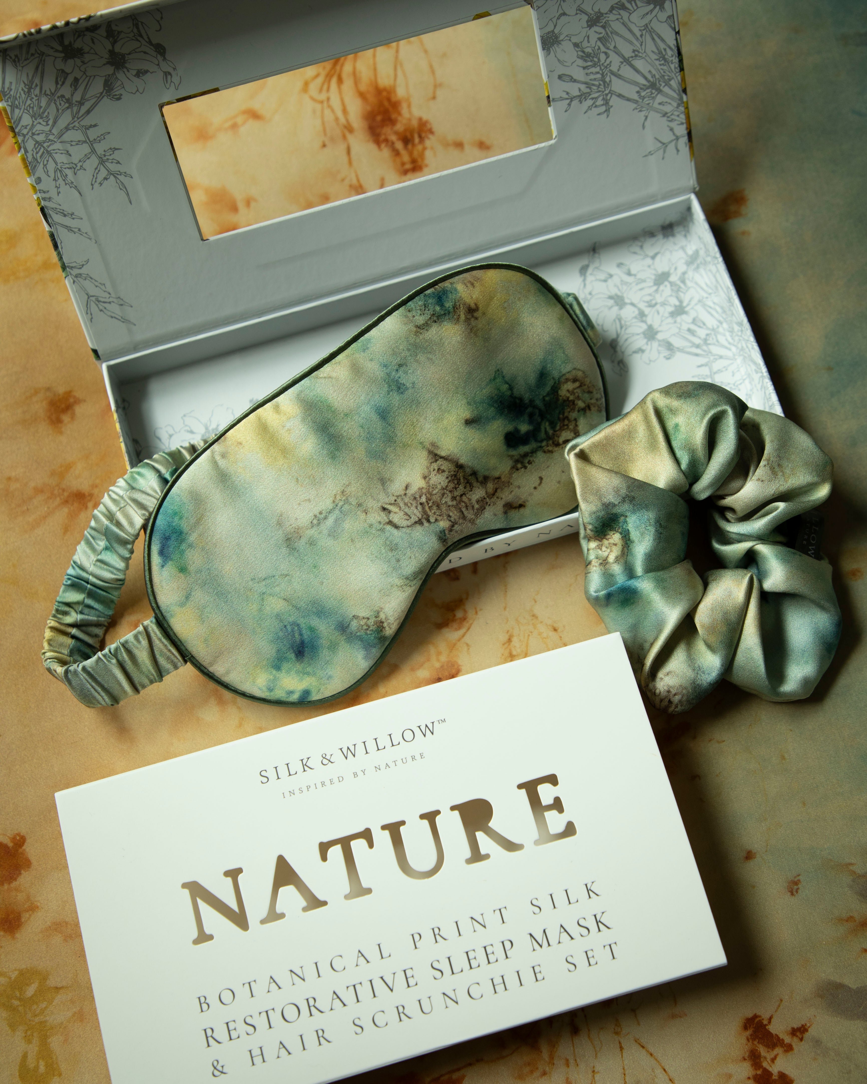 Silk & Willow Nature botanical print silk sleep mask and hair scrunchie set on a textured surface.