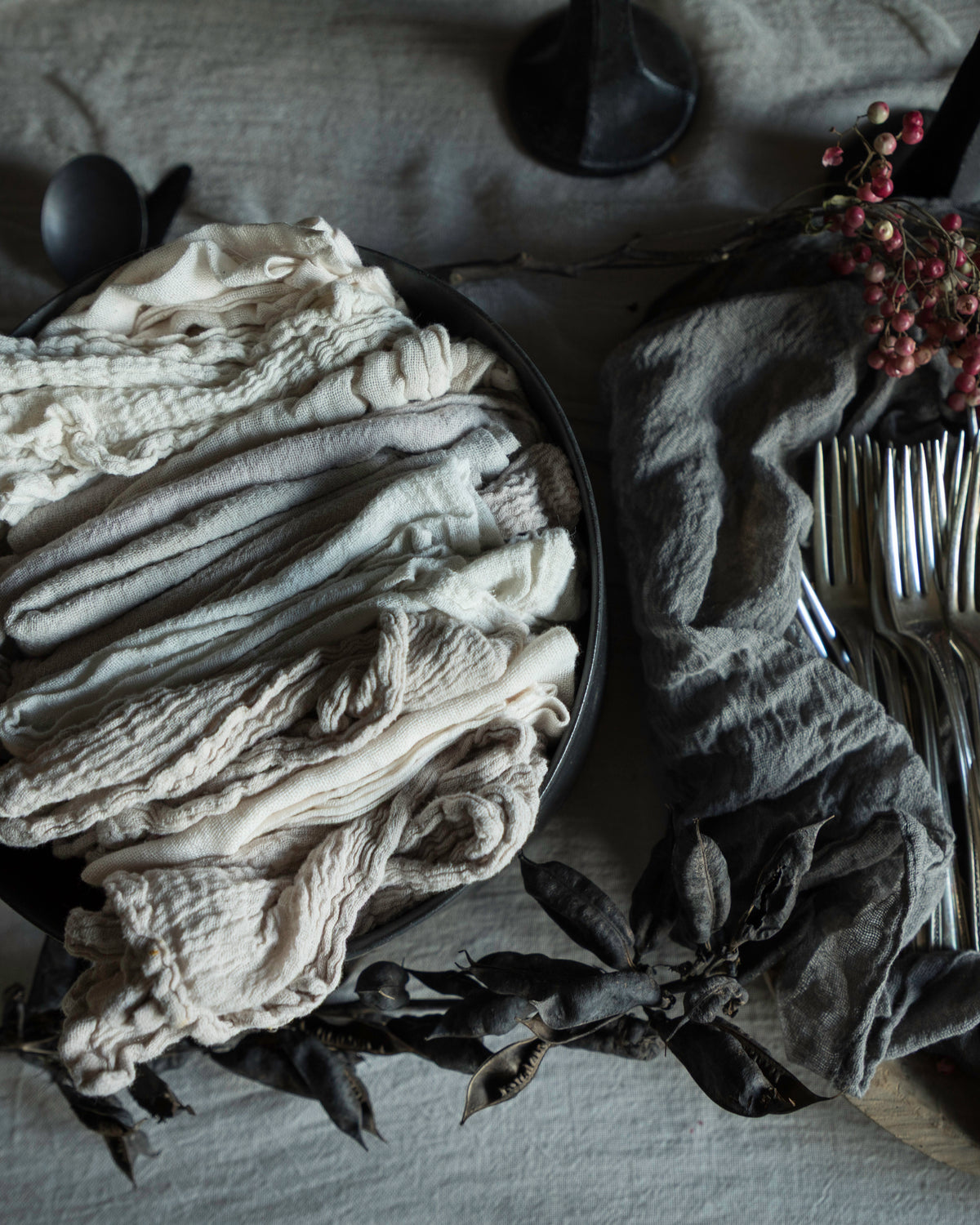 A styled flat-lay of folded fabric napkins or table linens in neutral tones, arranged alongside silver forks on a dark cloth with dried leaves and small red berries on a table covered in a light linen fabric.