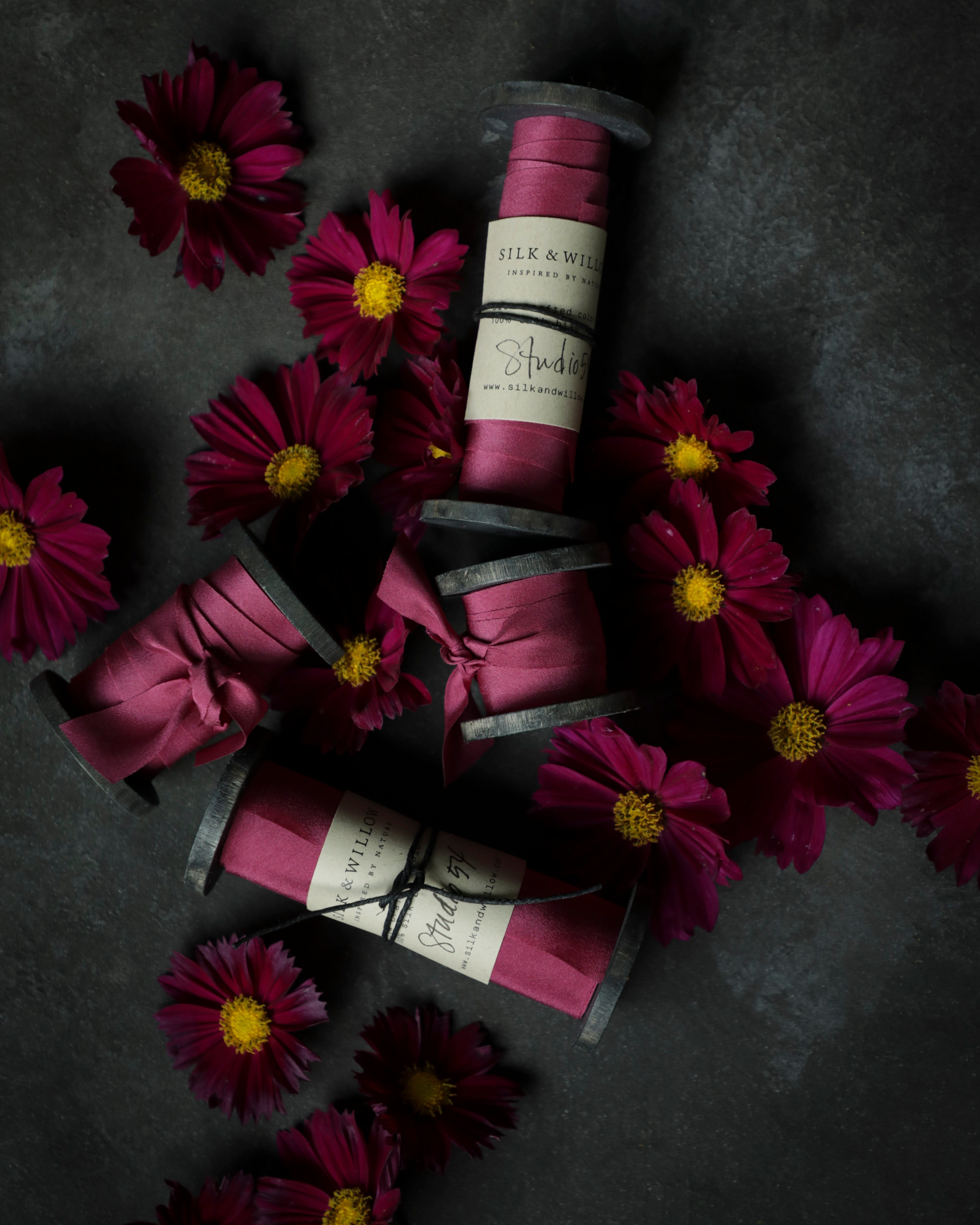 Spools of pink ribbon with labels on a dark surface surrounded by purple flowers.