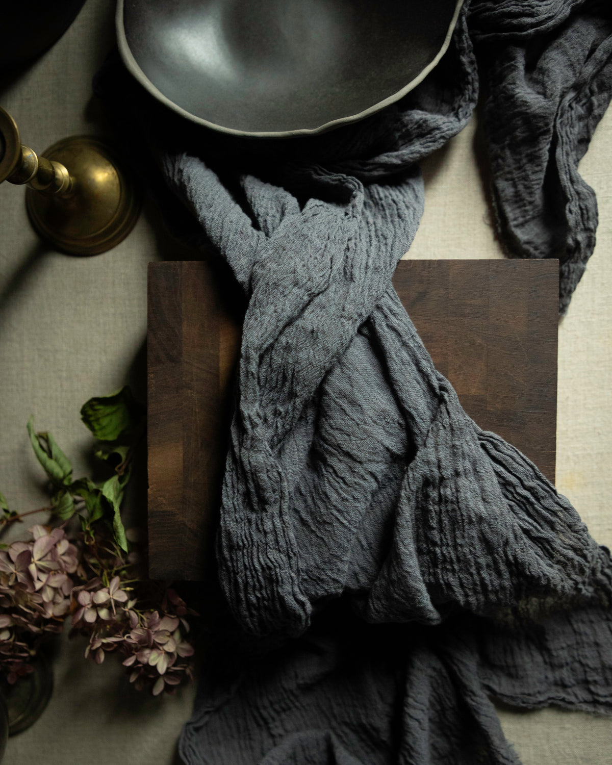 A dark charcoal gray crinkled gauze table runner draped over a dark wooden block, styled with a black bowl, a brass candle holder, and dried flowers on a neutral fabric background.