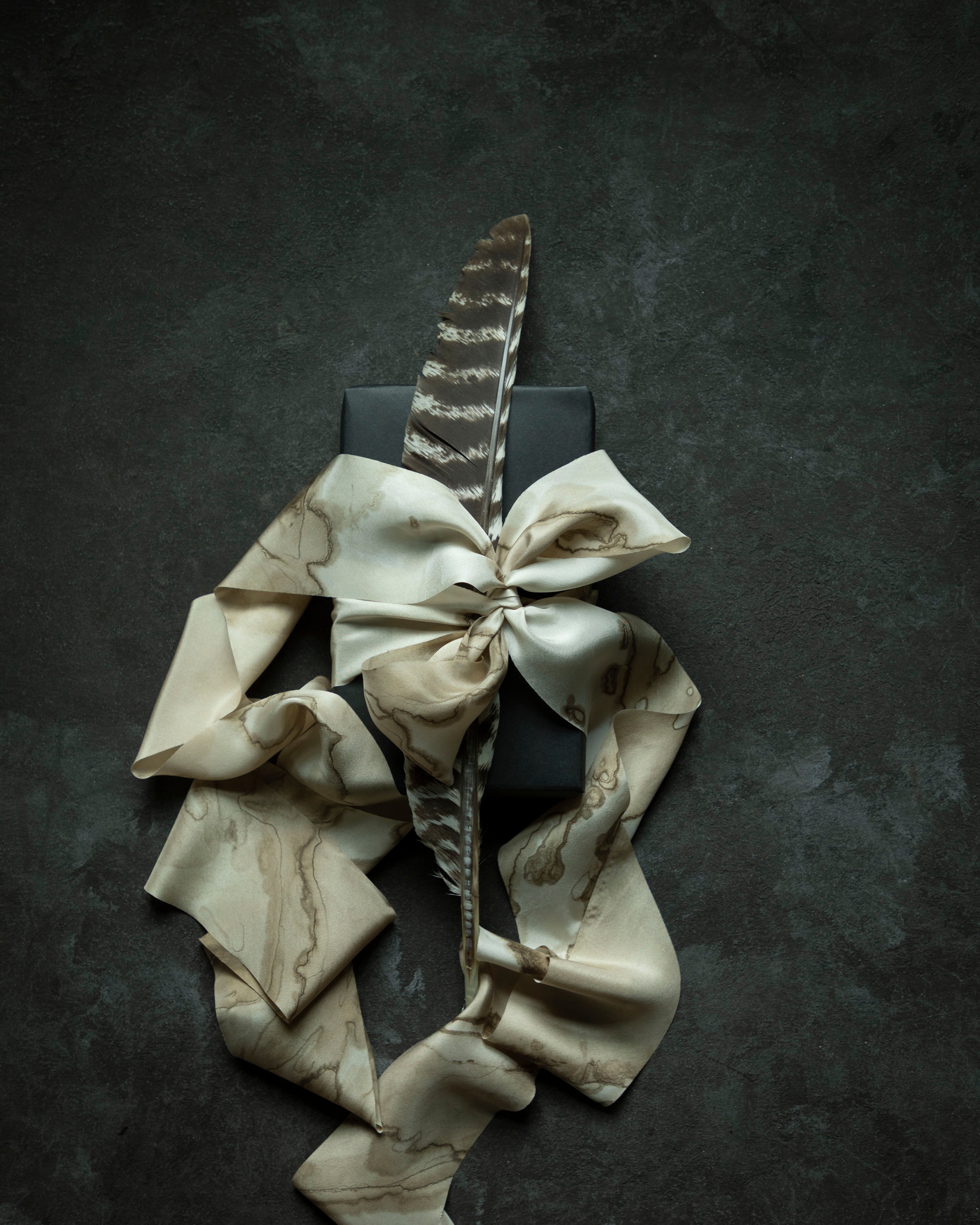 A cream and beige silk ribbon with a woodgrain marbled pattern, styled with a large bow and a single brown feather on top of a black rectangular box, set against a dark textured background.