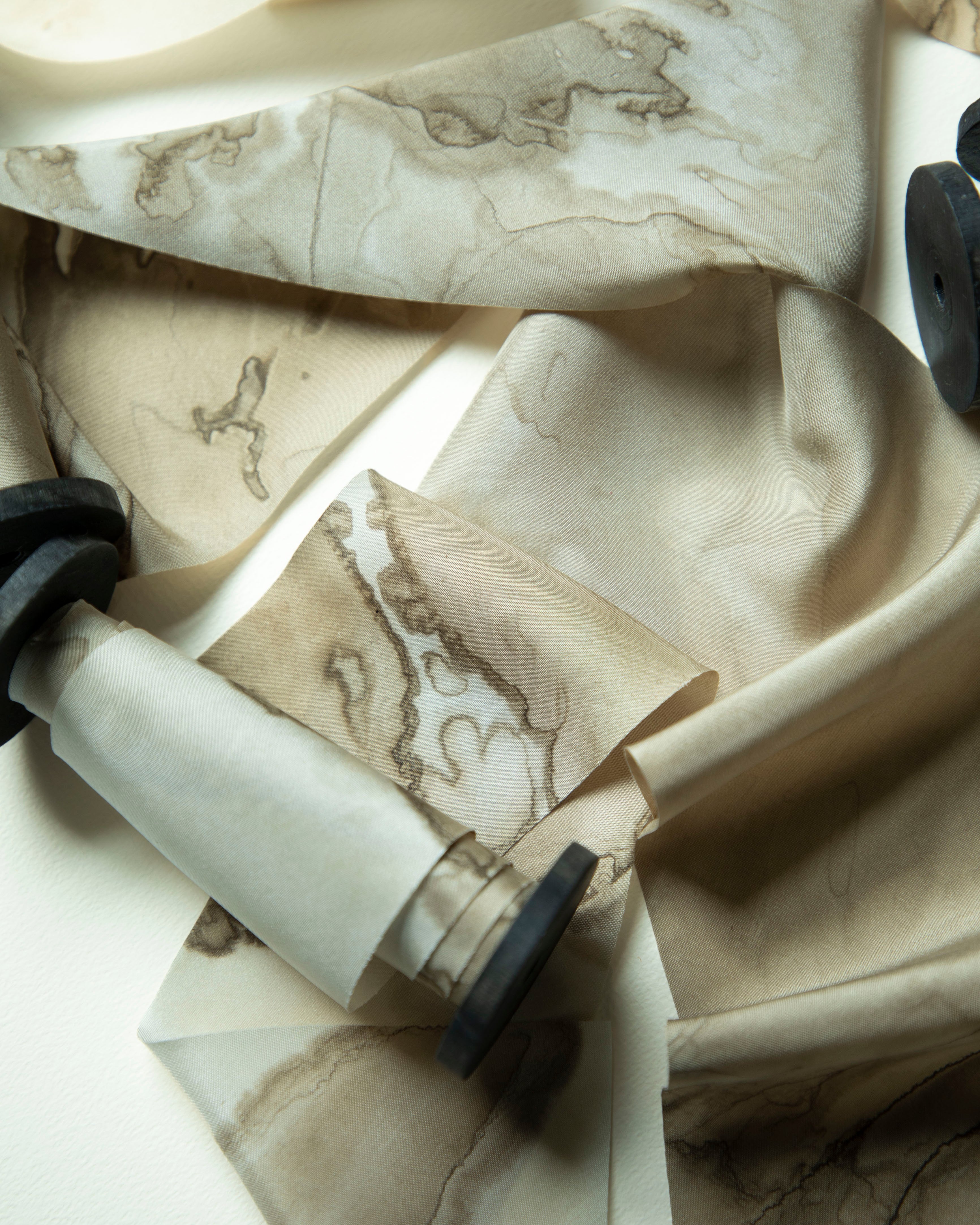 A close-up of draped silk ribbon with a beige and cream marble or stone-like pattern, arranged on a light surface with a few dark round props partially visible.