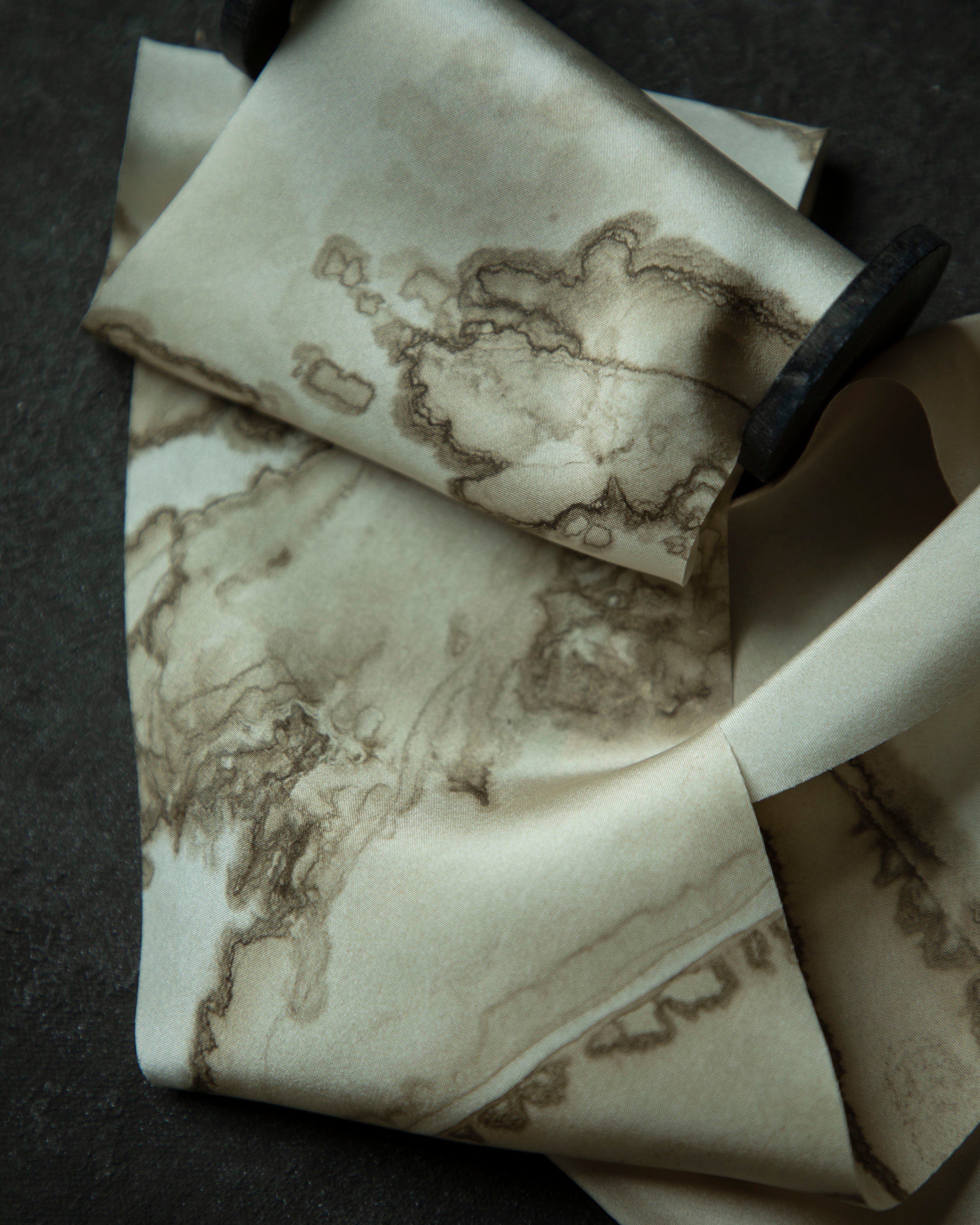 A close-up of a cream-colored silk ribbon with a brown abstract map or ink-blot pattern on a wood spool on a dark textured surface.