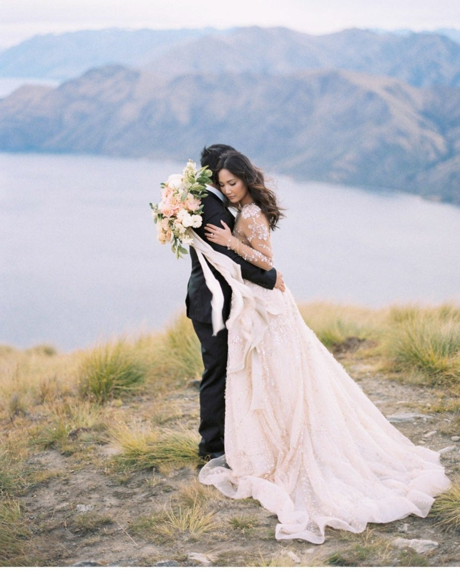 bride and groom hugging on New Zealand ridge with bridal bouquet and flowing silk and willow ribbon