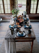 A rustic wooden dining table set with blue and gray ceramic dishes, dark mugs, glassware, and a draped gray table runner, placed in a sunlit cabin interior with large wood-framed windows and an outdoor view.