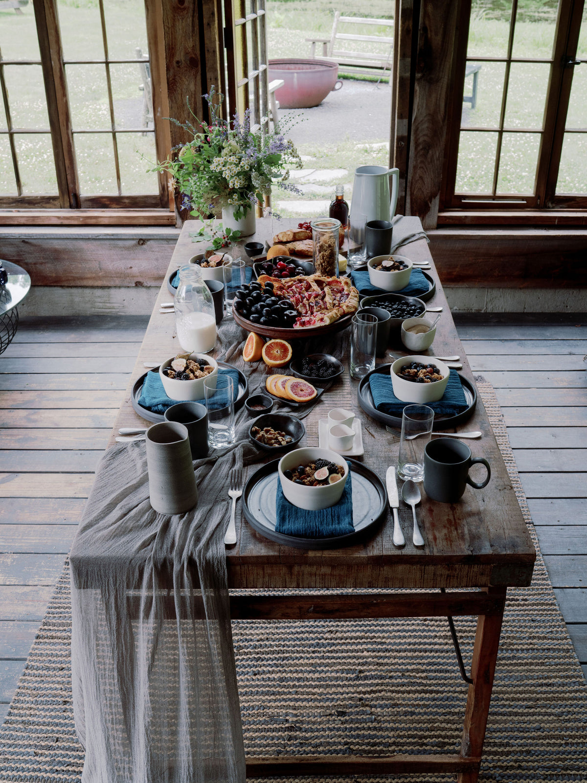 A rustic wooden dining table set with blue and gray ceramic dishes, dark mugs, glassware, and a draped gray table runner, placed in a sunlit cabin interior with large wood-framed windows and an outdoor view.