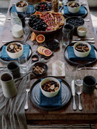 A rustic wooden dining table set for breakfast with bowls of cereal, blue napkins, plates, mugs, glasses, sliced fruit, and a large fruit and pastry platter in the center.