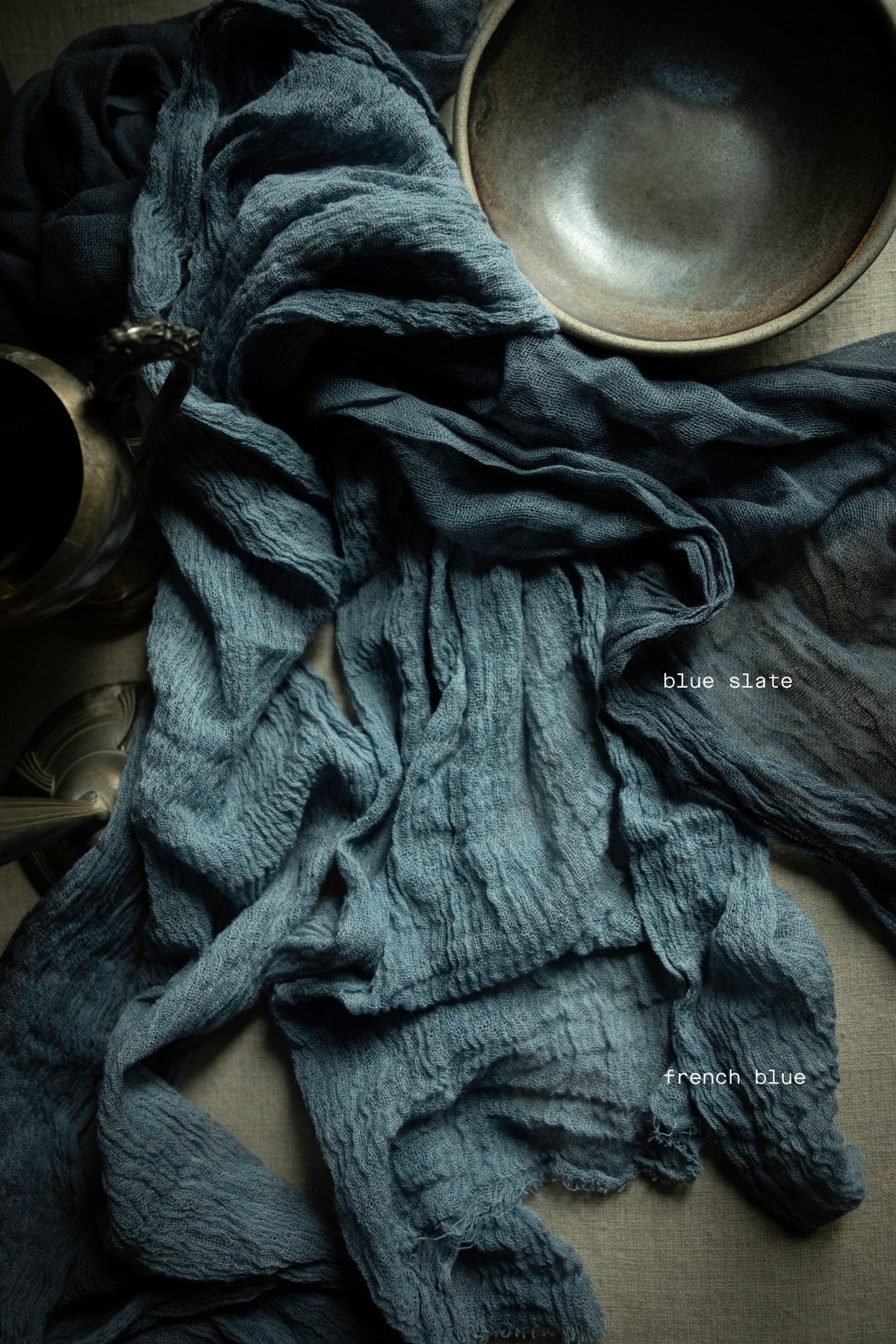 A crinkled dark blue-gray fabric, likely a scarf or textile, draped across a neutral surface with a ceramic bowl and metal vessel as props. Small text labels read 'Blue slate' and 'French blue'.