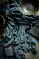 A crinkled dark blue-gray fabric, likely a scarf or textile, draped across a neutral surface with a ceramic bowl and metal vessel as props. Small text labels read 'Blue slate' and 'French blue'.