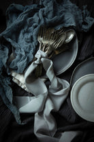 A styled flat-lay of silver cutlery tied with a white ribbon, set on dark gray plates and draped with a slate-blue textured cloth over a dark charcoal fabric background.