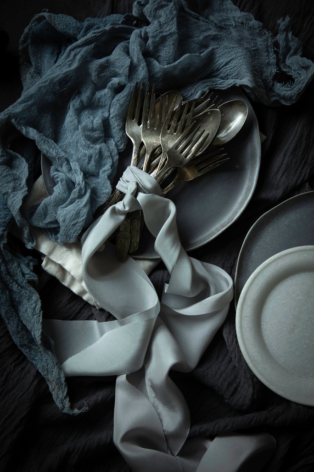 A styled flat-lay of silver cutlery tied with a white ribbon, set on dark gray plates and draped with a slate-blue textured cloth over a dark charcoal fabric background.