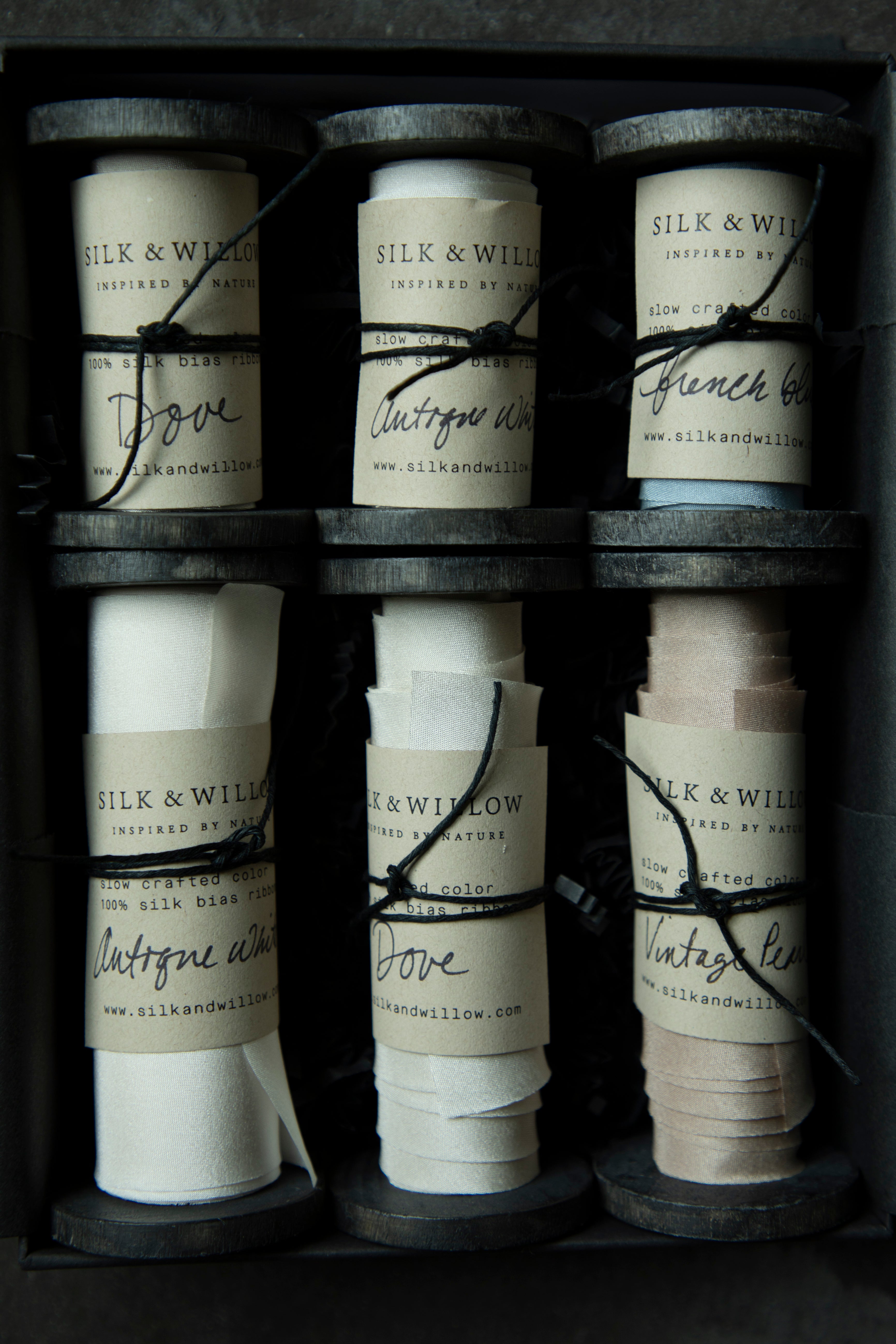 Six rolls of silk ribbon in different colors and widths arranged in a dark gift box. Each roll has a paper label with the brand name 'SILK & WILLOW' and a black string tied around it.