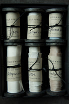 Six rolls of silk ribbon in different colors and widths arranged in a dark gift box. Each roll has a paper label with the brand name 'SILK & WILLOW' and a black string tied around it.