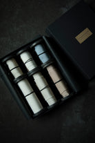 A luxury gift set of six premium ribbons in a black presentation box, shown from a top-down angle on a dark textured surface. The ribbons come in white, light blue, and light beige colors, with a gold-toned brand plaque on the lid.