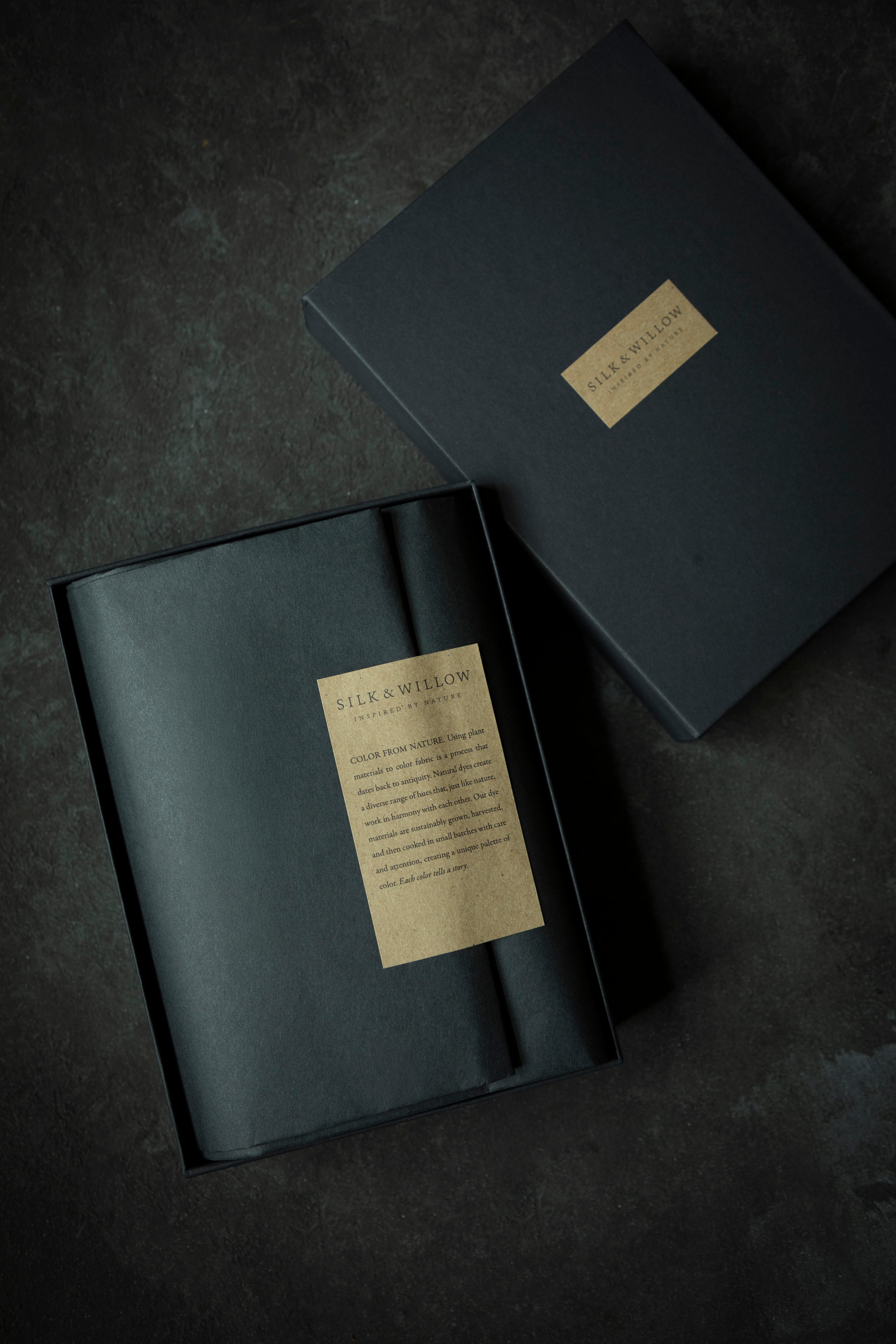 A dark charcoal gray presentation box with a gold label reading 'SILK & WILLOW' and 'The Art of Packaging', shown open with its lid placed beside it on a dark textured background.