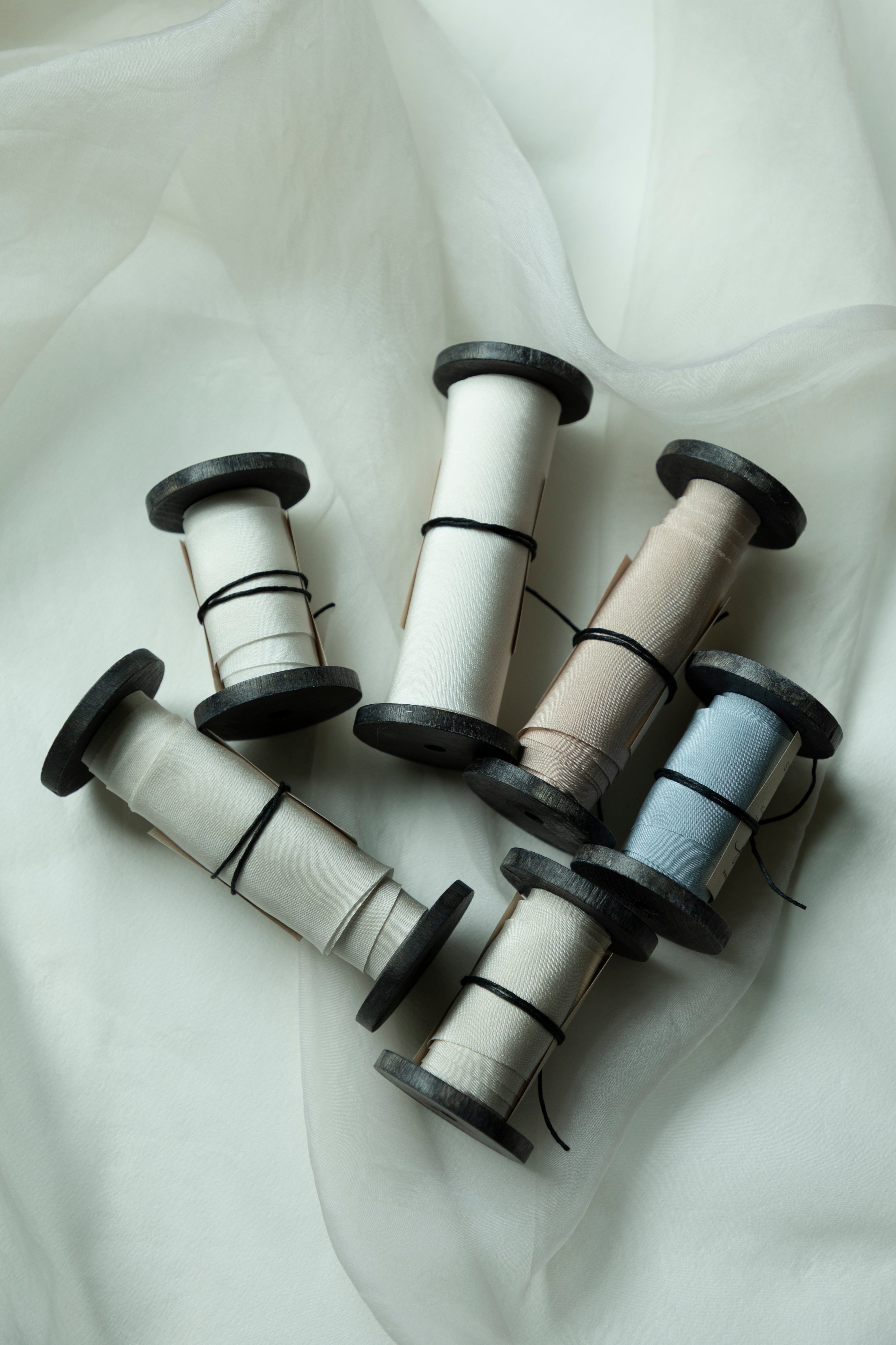Five spools of bridal bouquet silk ribbon in neutral and pastel shades, including cream, beige, light gray, and pale blue, arranged on a draped white fabric background.