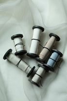 Five spools of bridal bouquet silk ribbon in neutral and pastel shades, including cream, beige, light gray, and pale blue, arranged on a draped white fabric background.