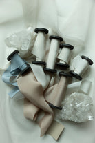 A styled flat-lay of multiple spools of hand-dyed silk ribbon in neutral tones, including beige, cream, light blue, and taupe, arranged on a draped white fabric background with clear crystal clusters.
