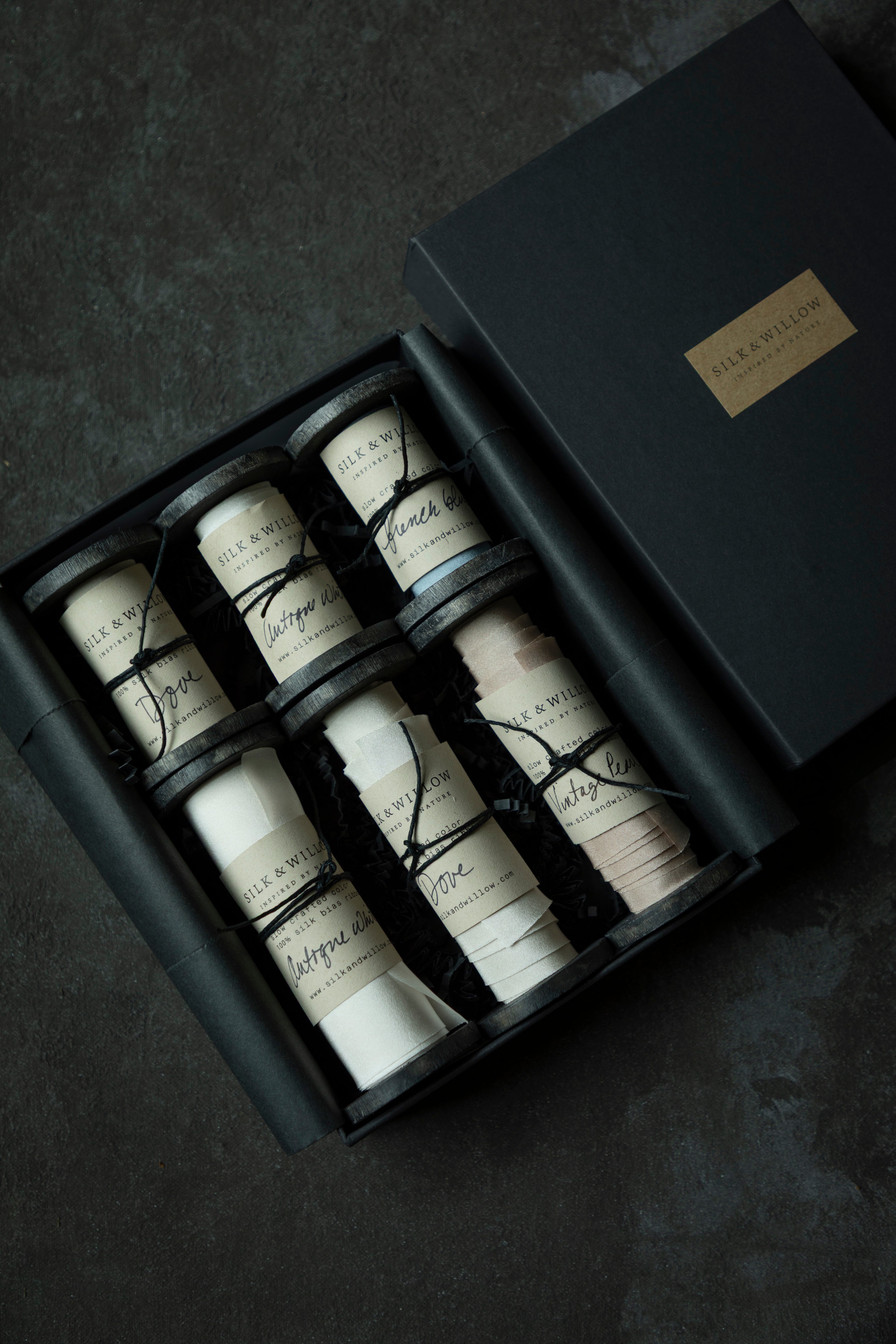 An open black gift box containing six spools of premium silk ribbon, shown on a dark textured surface. The closed matching box with a gold label sits beside it.