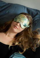 A woman wearing a botanical print silk sleep mask is lying on a blue pillow.