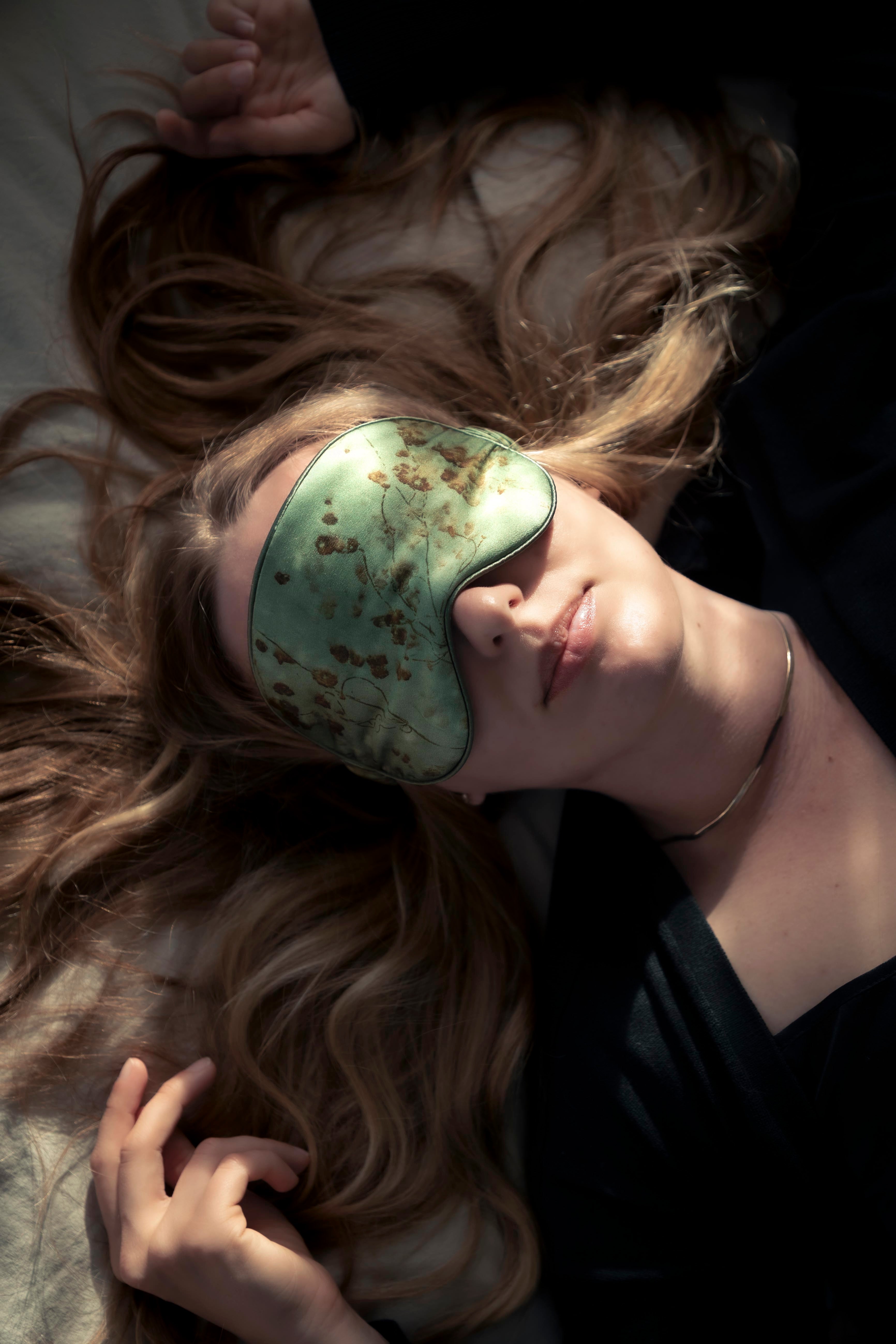a woman lying on a bed in the sunlight wearing a botanical print organic silk sleep eye mask 