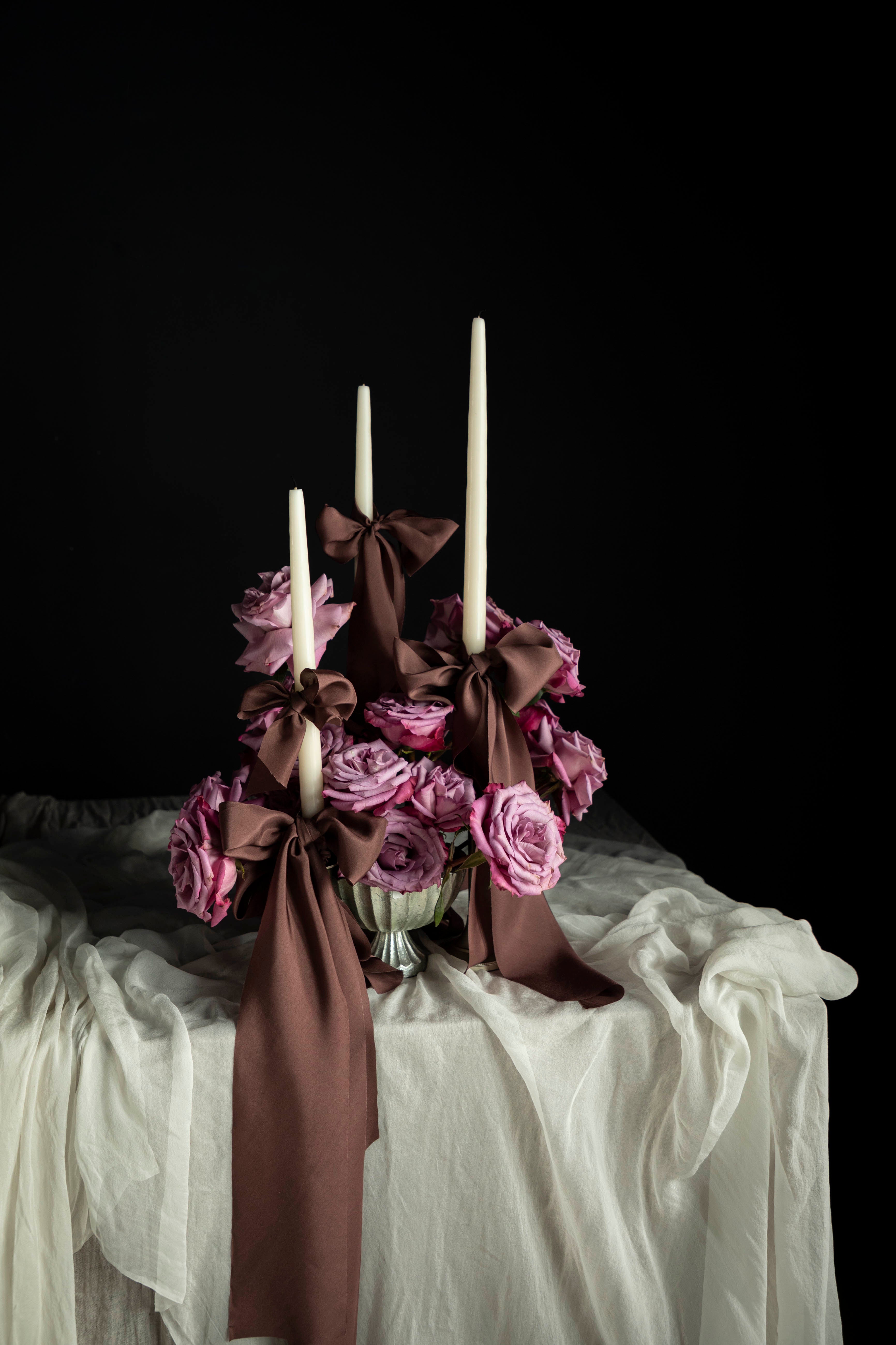 Decorative floral arrangement with candles on a white surface against a black background. featuring brown silk ribbon and mauve roses.