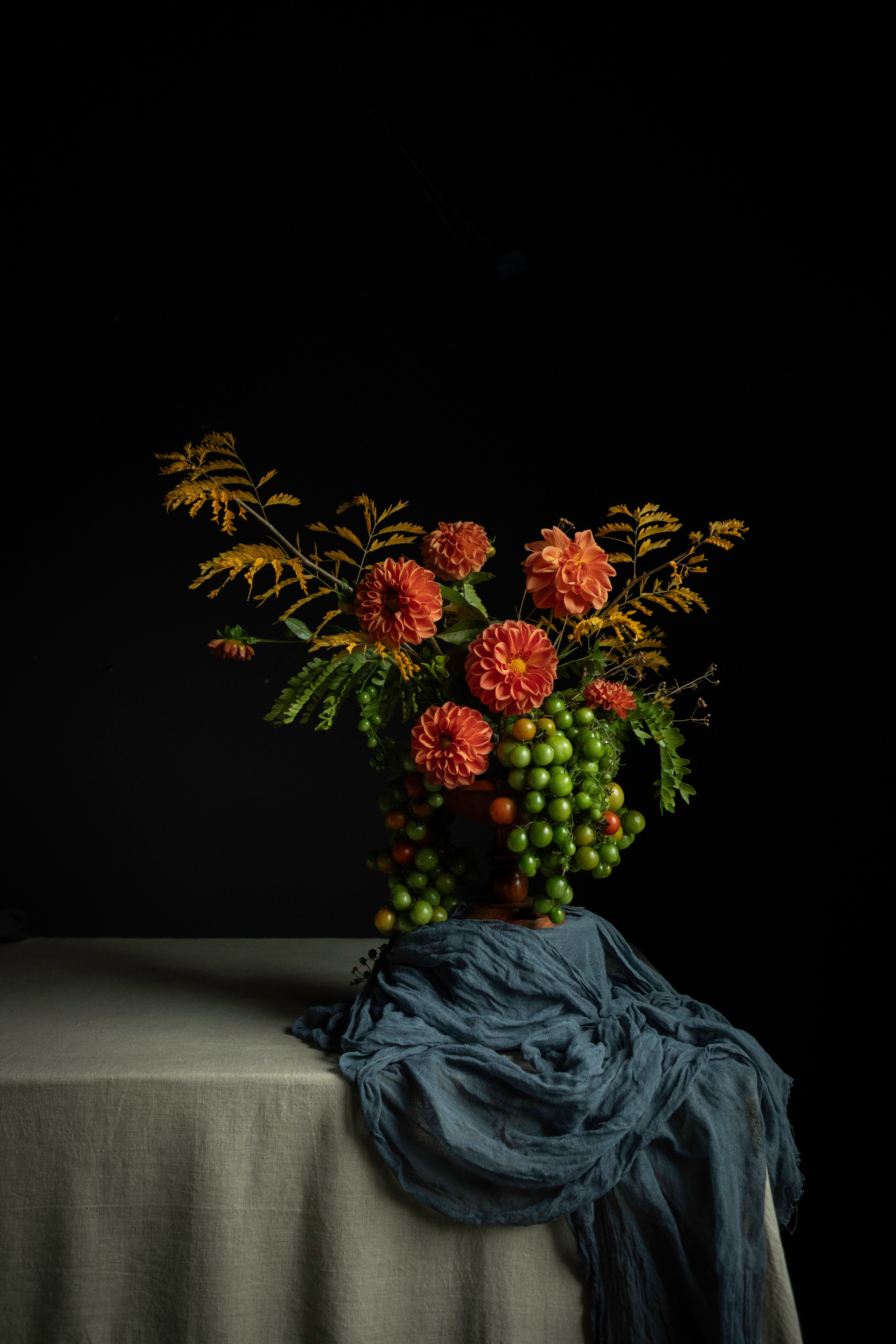 A still life floral arrangement with orange flowers, green leaves, and clusters of green and red berries, set on a draped table against a black background.